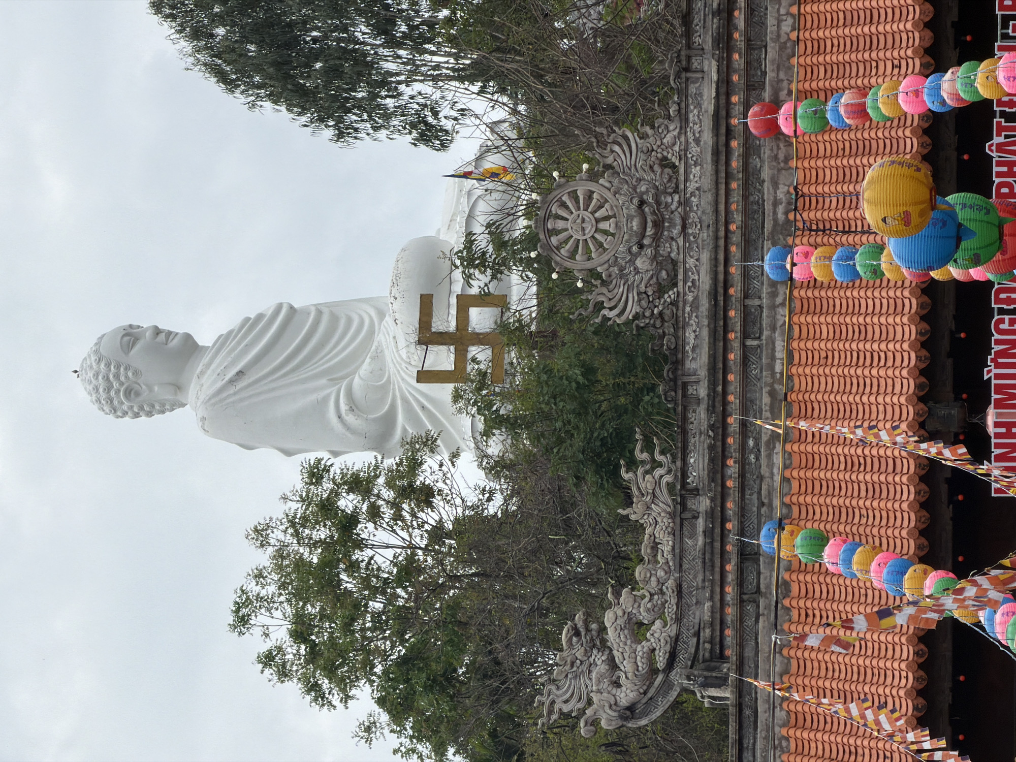 The image captures a large, white reclining Buddha statue in Tay Nha Trang Ward, Vietnam. The statue is positioned outdoors against a cloudy sky and surrounded by trees and shrubbery. In front of the statue are religious symbols, including a swastika and a dharma wheel. A building with an orange tile roof is partially visible, adorned with colorful lanterns and Buddhist prayer flags. The building also features ornate stone carvings.