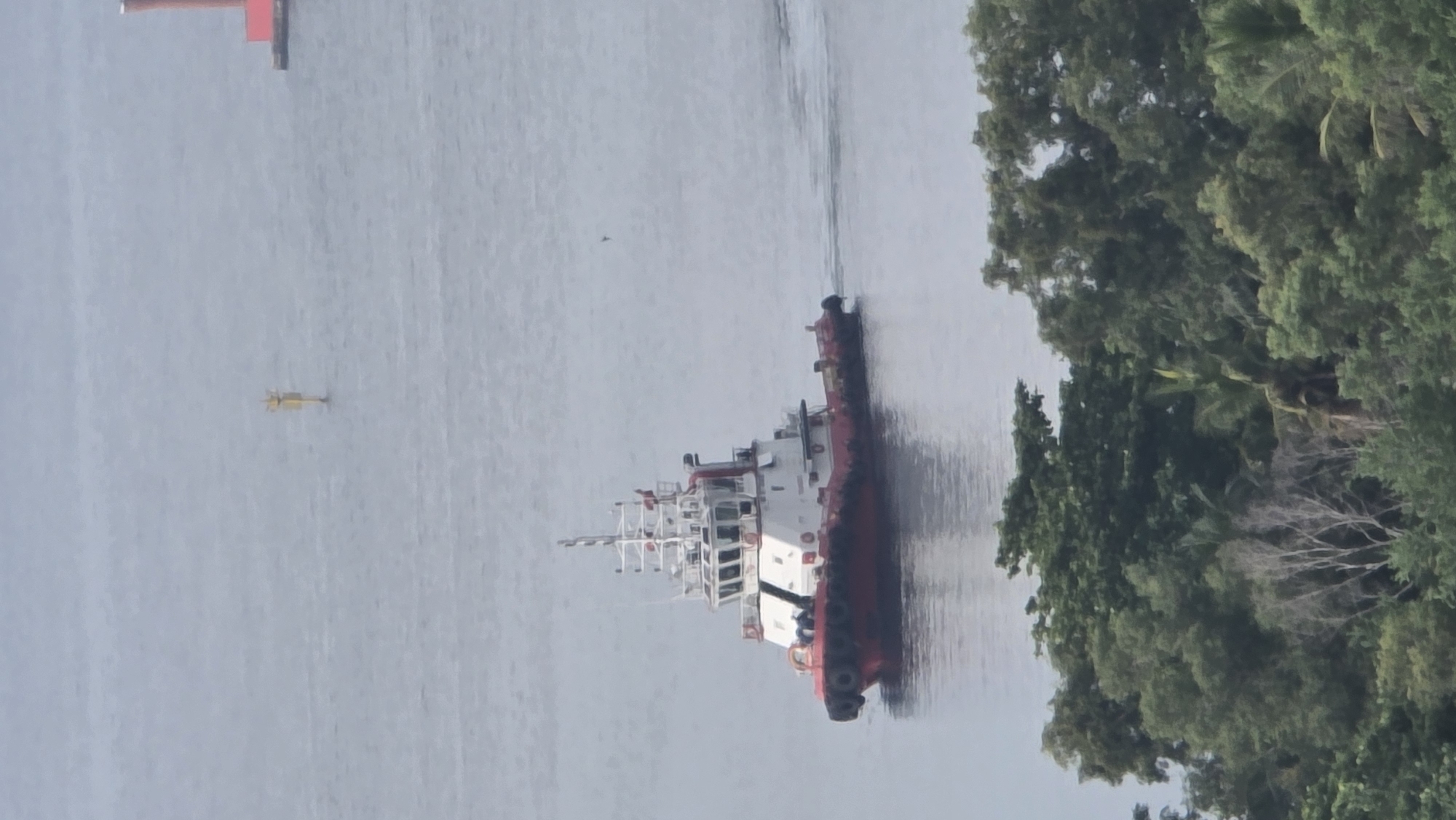 The image shows a red and white workboat navigating a waterway. To the boat's left, in the distance, is a smaller yellow object that could be a buoy or small boat. Lush greenery flanks the right side of the waterway, suggesting a coastal or river environment. In the far distance, a red structure sits on the shoreline. Given the mention of Lhokseumawe, Indonesia, it is likely that this scene is of a coastal area near or within the city, perhaps a harbor or river leading to the sea. The day appears overcast, resulting in subdued lighting and a grey tone in the water.