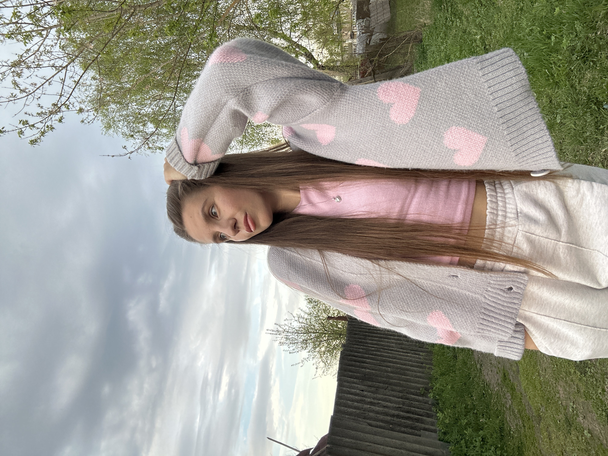 A young woman is the central subject of the image, posed outdoors against a backdrop of sky, trees, and vegetation. She is wearing a pink top, light-colored sweatpants, and a gray cardigan adorned with pink hearts. Her long, straight hair is loose around her shoulders. Her hand is raised near her head. The background includes trees, a cloudy sky, and what appears to be a corrugated roofing material in the lower part of the frame. The location is identified as Борщевка Малая, Russia.