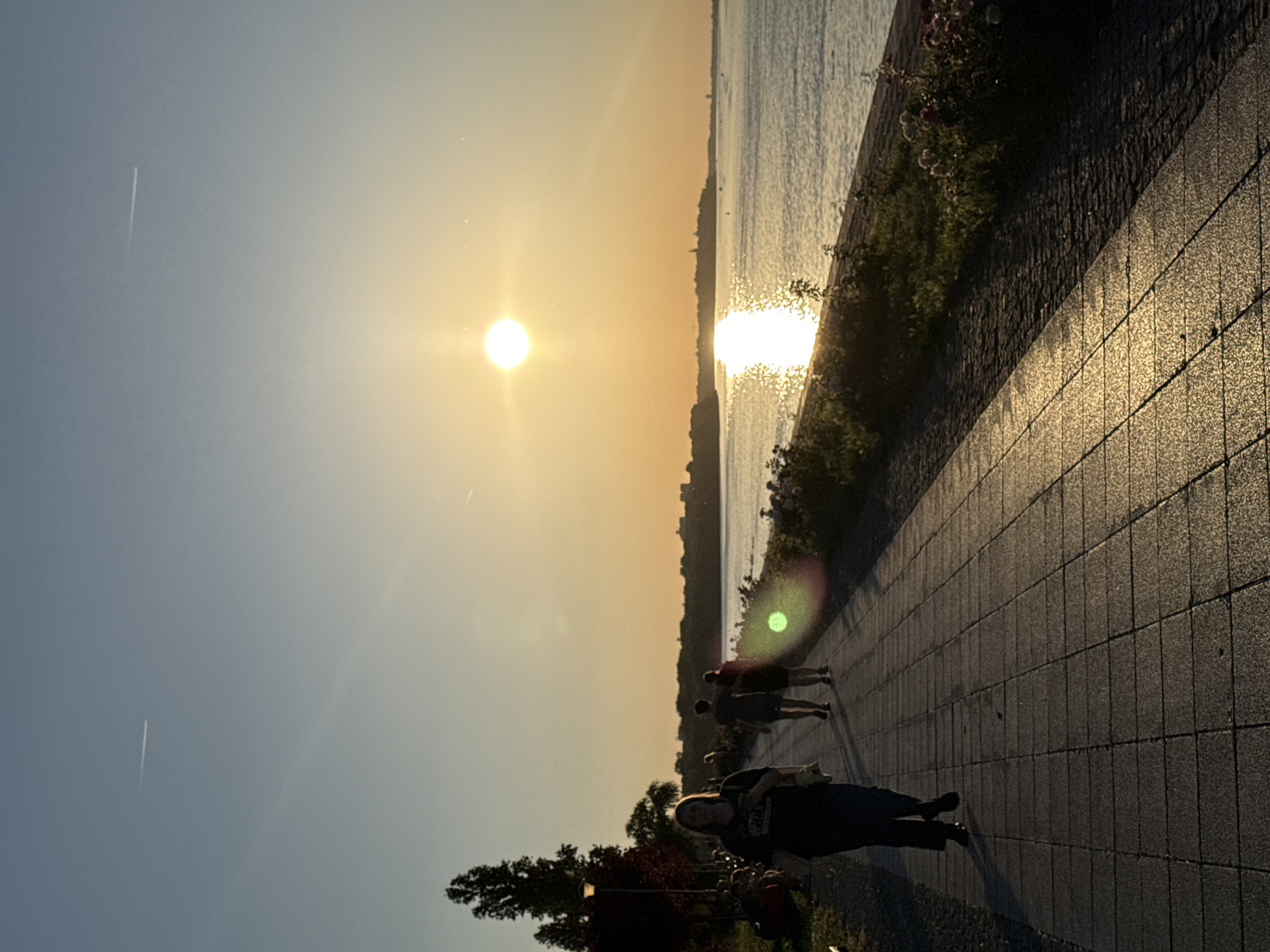 The image shows a riverside scene in Belgrade, Serbia, likely along the Sava or Danube River. The sun is setting, casting a bright glow on the water and creating long shadows. A paved walkway runs along the river, with greenery and flowers lining the edge. Several people are walking along the walkway, their silhouettes visible against the bright light. In the sky, a contrail from an airplane can be seen.