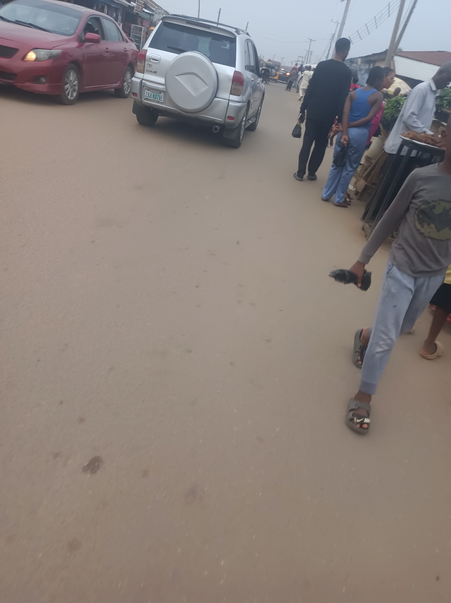 The image depicts a street in Narayi, Nigeria, on what appears to be a typical day, possibly a market day. There is a red sedan and a silver SUV with license plate ZAR-447AE. Several people are walking along the side of the road. A boy in a gray shirt and light blue pants is walking toward the viewer holding a black object in his hand. Some of the individuals in the background appear to be engaged in commercial activity along the roadside. The weather is overcast.