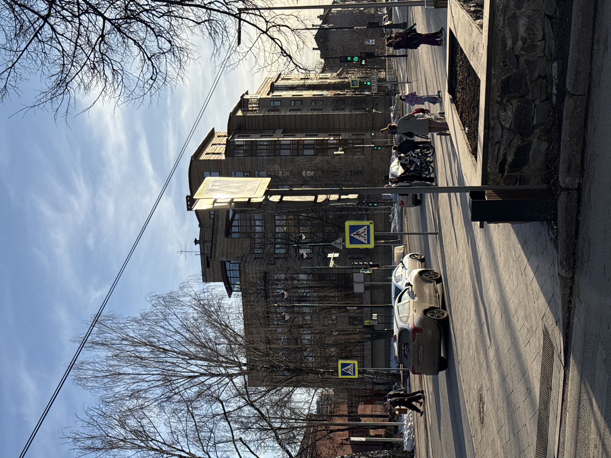 An urban street scene unfolds in Perm, Russia, at the intersection of Ulitsa Gazety Zvezda and Komsomolsky Prospekt, with the distinctive multi-story residential building at Ulitsa Gazety Zvezda, 12 visible prominently in the background. The time is daytime, characterized by bright sunlight from a low angle, casting long shadows across the paved roads and sidewalks. The sky is clear with scattered wispy clouds.
The environment shows bare deciduous trees, indicating a late winter or early spring setting, with patches of snow and ice remaining on the ground, particularly near sidewalks and tree bases. Overhead utility lines traverse the upper portion of the frame.
Vehicles include a light brown or beige SUV, possibly a Renault Arkana, with a small object on its roof (potentially a taxi light), positioned in the left lane of the main road, facing away from the viewer. Behind it, in the right lane, is a silver sedan, also moving away. Additional vehicles are visible further down the street.
Approximately 10-12 individuals are present, primarily pedestrians. Two adult males in dark clothing walk from left to right on the foreground sidewalk. A woman in a light-colored coat pushes a baby stroller across a pedestrian crossing in the mid-ground. To the right, a child on a blue kick scooter, accompanied by two adults, walks towards the viewer on the sidewalk. Other pedestrians are visible on various sidewalks and crossing points throughout the scene.
Street furniture includes multiple yellow-bordered square signs indicating pedestrian crossings, traffic light poles displaying green signals for vehicular traffic, and a white "СТОП" (STOP) sign mounted on a pole. A stone-clad planter box filled with dark soil and a dark waste bin are visible on the right side of the foreground.