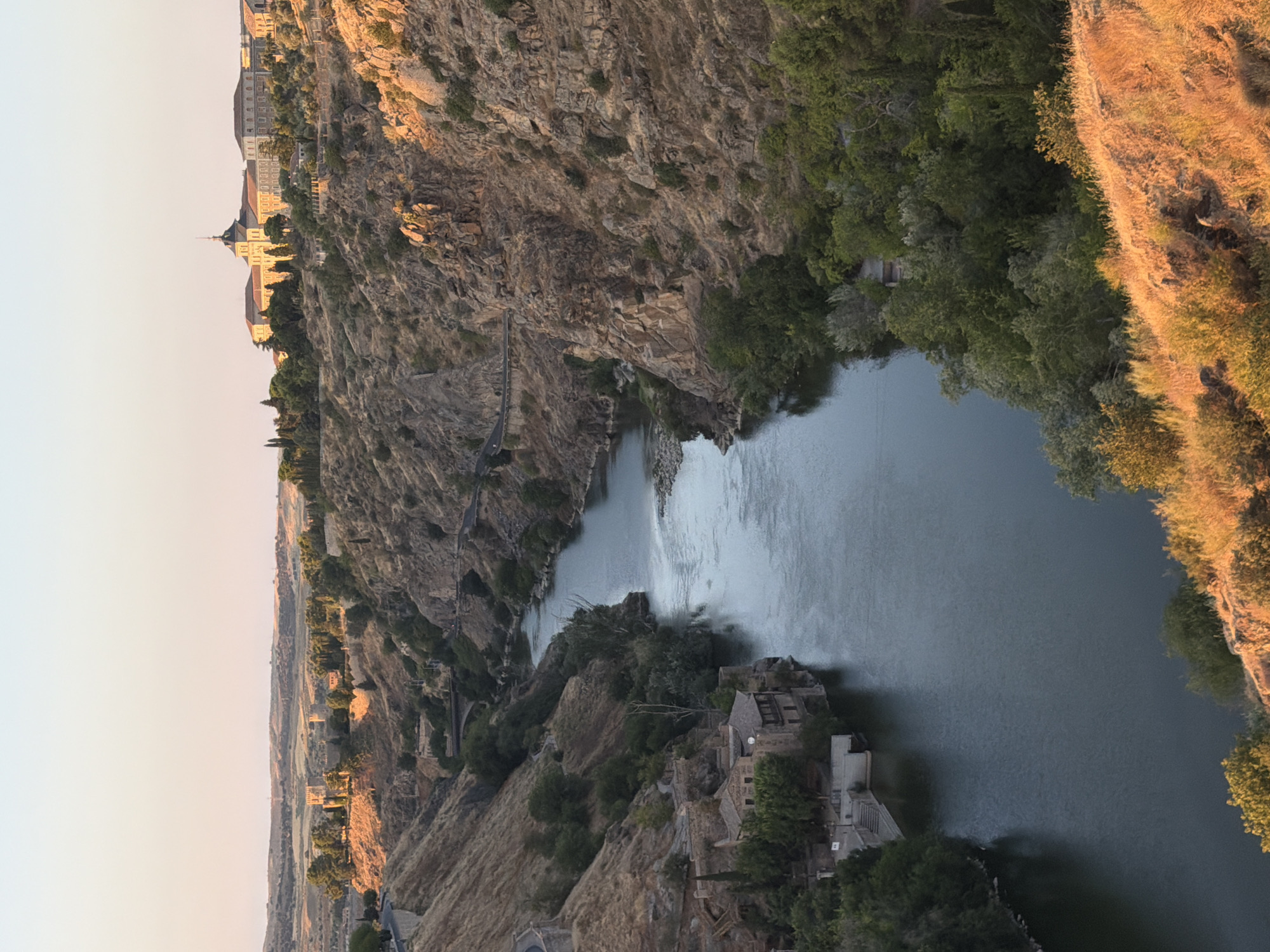 The image depicts a view of the Tagus River flowing through a gorge in Toledo, Spain. The sun is setting, casting a warm glow on the landscape. On the left bank, ancient stone buildings with tiled roofs are nestled into the hillside, some partially obscured by lush green trees. A road winds its way along the steep, dry, grassy slopes of the gorge. On the right bank, a rocky, arid cliff face rises, with a road visible traversing its upper section. In the distance, a large, light-colored building complex with multiple structures and a distinctive central tower sits atop a hill, surrounded by trees. The sky above is a soft gradient of pink and pale blue, indicative of twilight. The overall scene conveys a sense of historical depth and natural beauty.