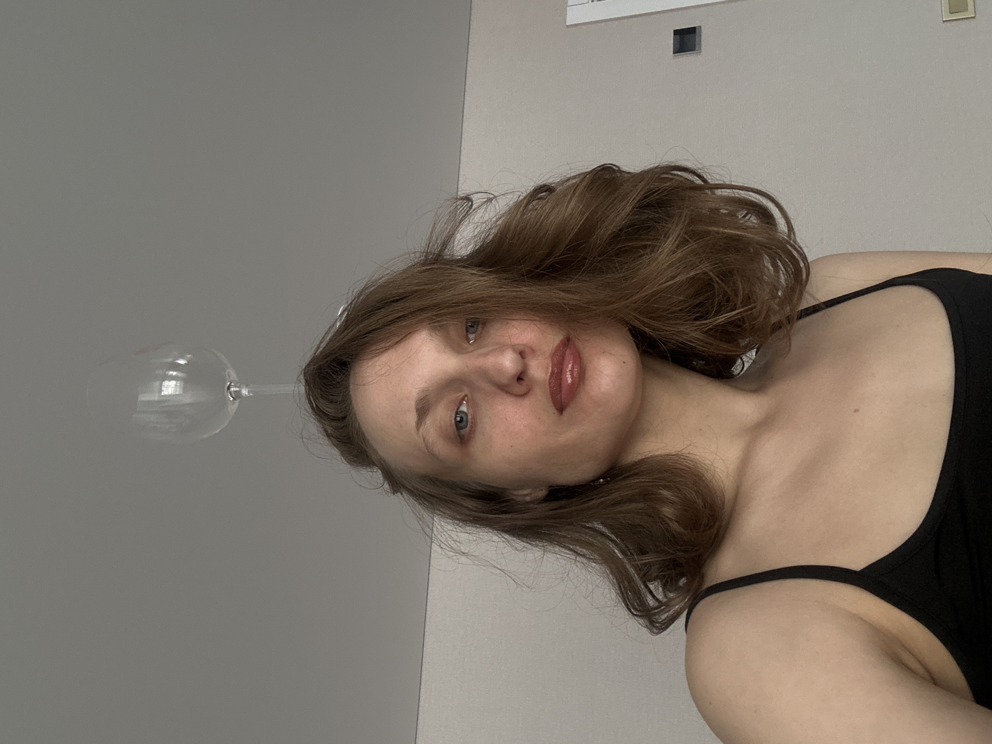 A young woman with fair skin and brown hair is posing for a photo. She is wearing a black top with thin straps. Her lips are glossy, and she appears to have light makeup on. In the upper left corner of the image, a clear wine glass is visible, positioned as if hovering next to her head. The background consists of two different shades of gray walls, with some small electrical fixtures near the top of the frame.
There is no information available to place the photograph in the city of Sepych, Russia.