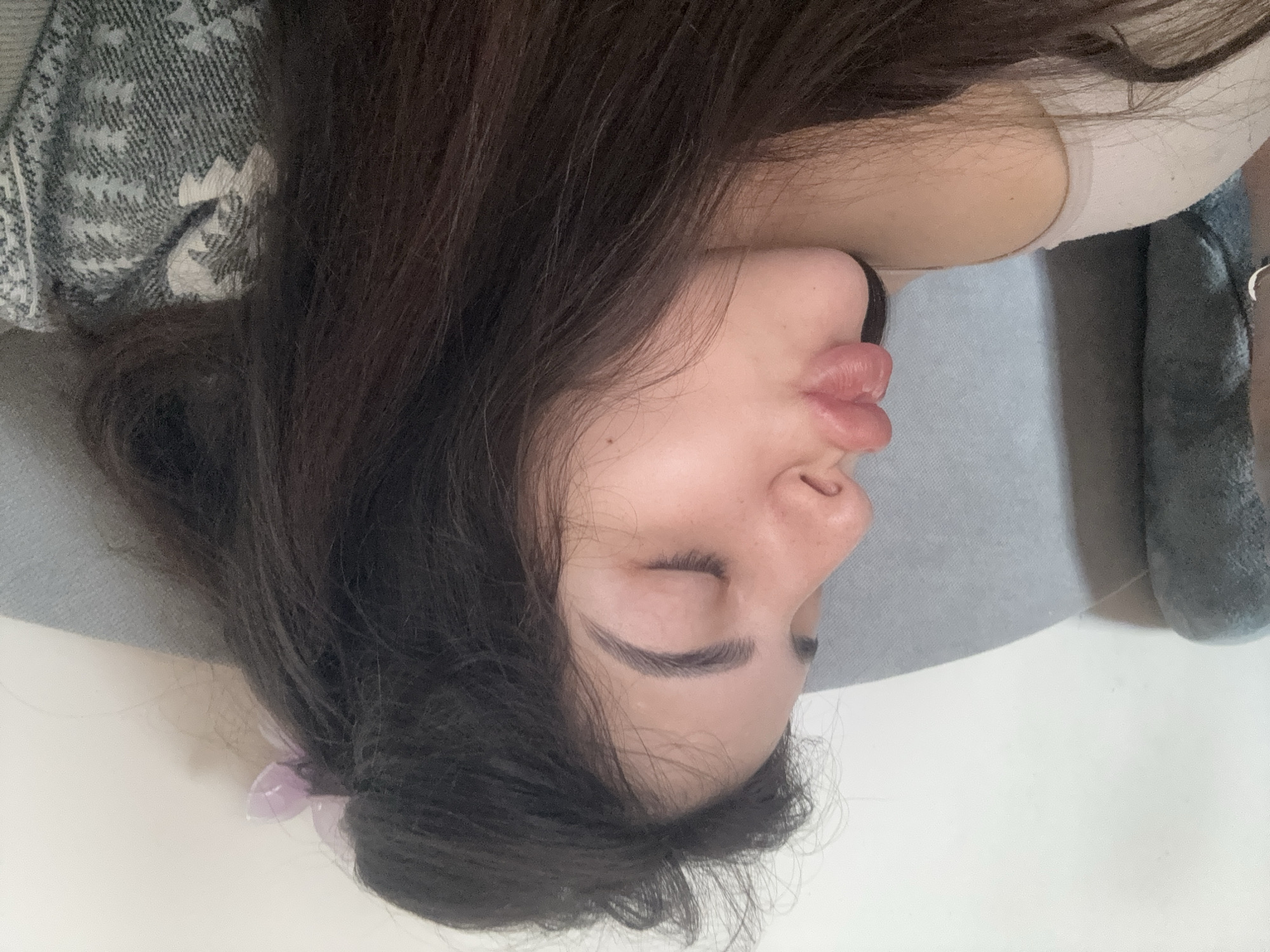 A close-up, horizontally-oriented photograph captures a young woman resting, possibly asleep or dozing. Her head is positioned on the left side of the frame, with her dark, long hair fanned around her face and over her shoulders. She is depicted in profile, with her eyes closed and her lips slightly pursed, suggesting a state of relaxation.
A light-colored top or bra strap is visible near her right shoulder. Her head rests on a light grey surface, likely a pillow or cushion. To the upper left, a patterned grey and white blanket or throw is partially visible, draped near her head. A small, translucent light purple hair accessory is nestled within her hair towards the bottom of the frame. On the far right, a dark grey object with a subtle metallic detail, possibly clothing or footwear, is partially visible. The lighting is soft and diffuse, indicating an indoor setting. There is no visible text. The image conveys a peaceful and serene atmosphere.