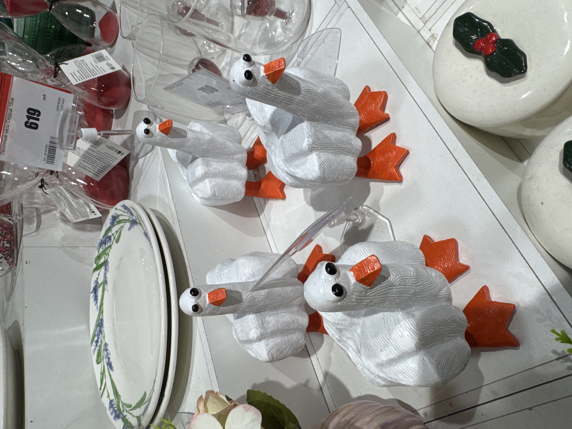 This image depicts a retail display shelf featuring four white, stylized goose figurines with orange beaks and feet. The figurines are arranged in a staggered formation, with two in the foreground and two behind them. To the left of the geese, a stack of white plates with a lavender floral pattern is visible. Behind the geese, an array of glass hourglasses filled with red sand and other assorted glassware are on display. A price tag indicating "619" with Russian text is attached to one of the hourglasses. The display appears to be set within a retail environment, possibly a home goods or gift shop. There are no people visible in the image, and no specific geographical markers for Ufa, Russia are present within the frame.