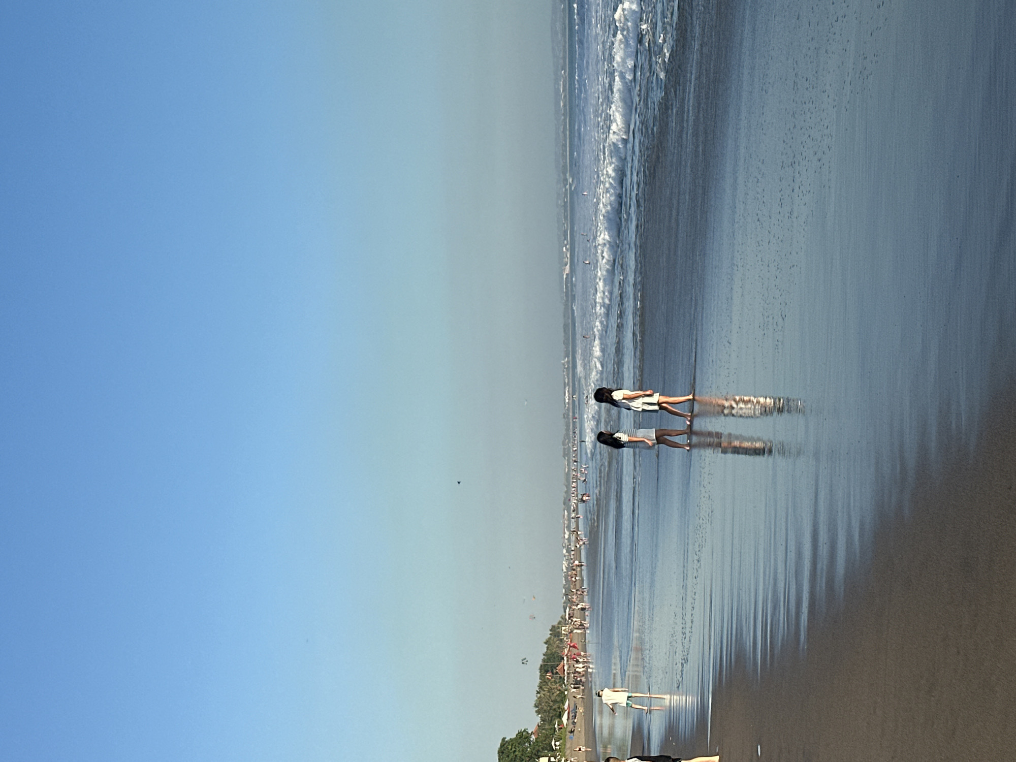 This media file depicts a wide, sandy beach with gentle waves lapping at the shore. The scene is bathed in the bright, clear light of a sunny day, suggesting it is mid-morning or early afternoon. The sky is a vibrant, cloudless blue, transitioning to a paler hue closer to the horizon.
Several people are visible on the beach. In the foreground, two individuals, likely a child and an adult, are walking along the wet sand, their reflections clearly visible in the shallow water. Further down the beach, more people can be seen walking and interacting. In the middle distance, there are several small boats or surfboards in the water, and some kites are flying in the air, adding a sense of activity and recreation to the scene. The background shows a line of palm trees and some structures, indicating a tropical location.
The overall mood of the image is one of peaceful leisure and enjoyment of a beautiful day at the beach. The reflections in the water and the clear sky contribute to a sense of tranquility. There are no visible text elements in the media file. Given the location context of North Kuta, Indonesia, this scene likely captures a typical day at a popular tourist beach.
