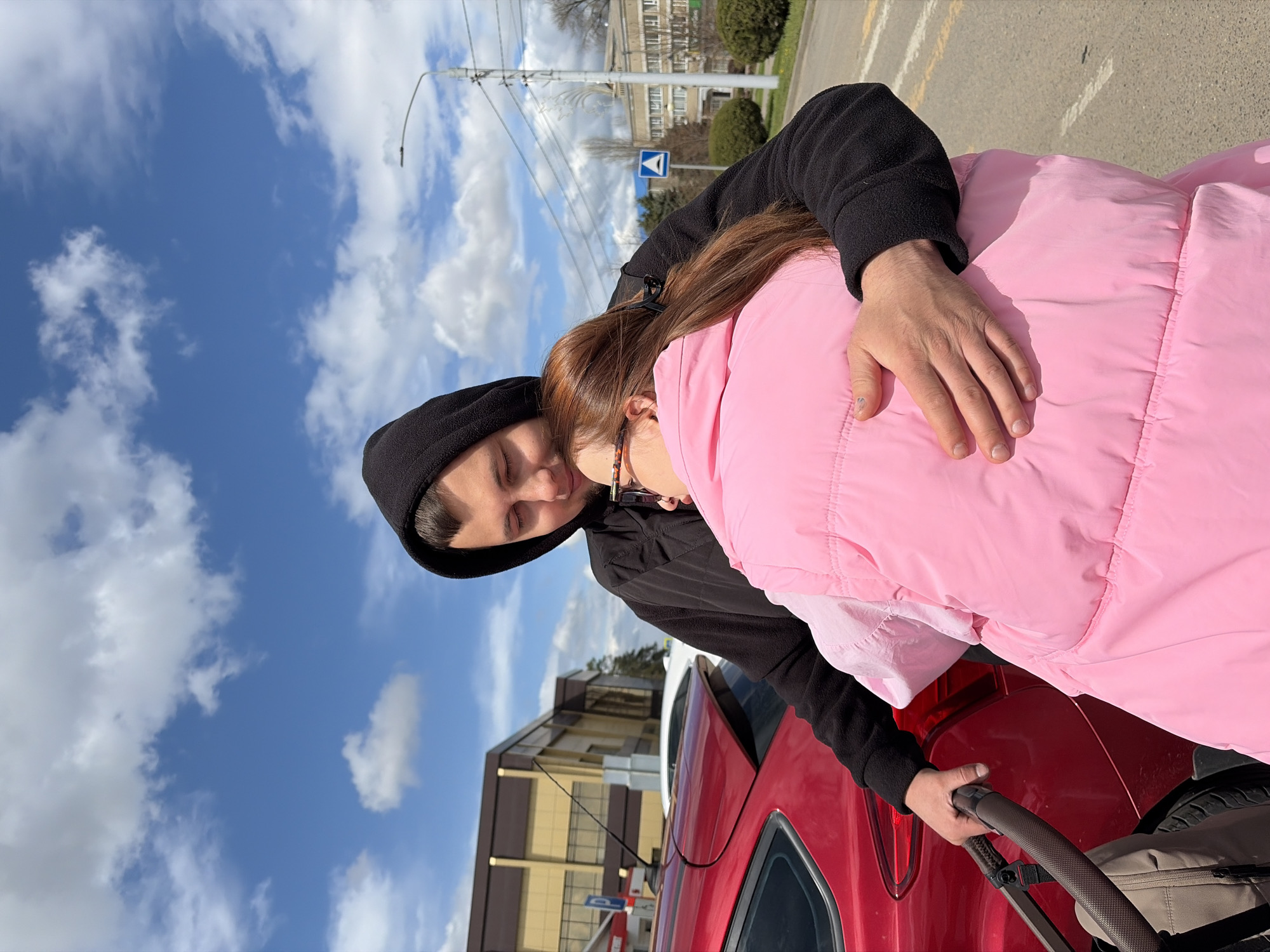 A man wearing a black hoodie and a woman in a pink jacket are hugging in front of a red car and a baby stroller. The location is Novopokrovskaya, Russia, as suggested by the surrounding buildings and infrastructure. The sky is blue with white clouds.