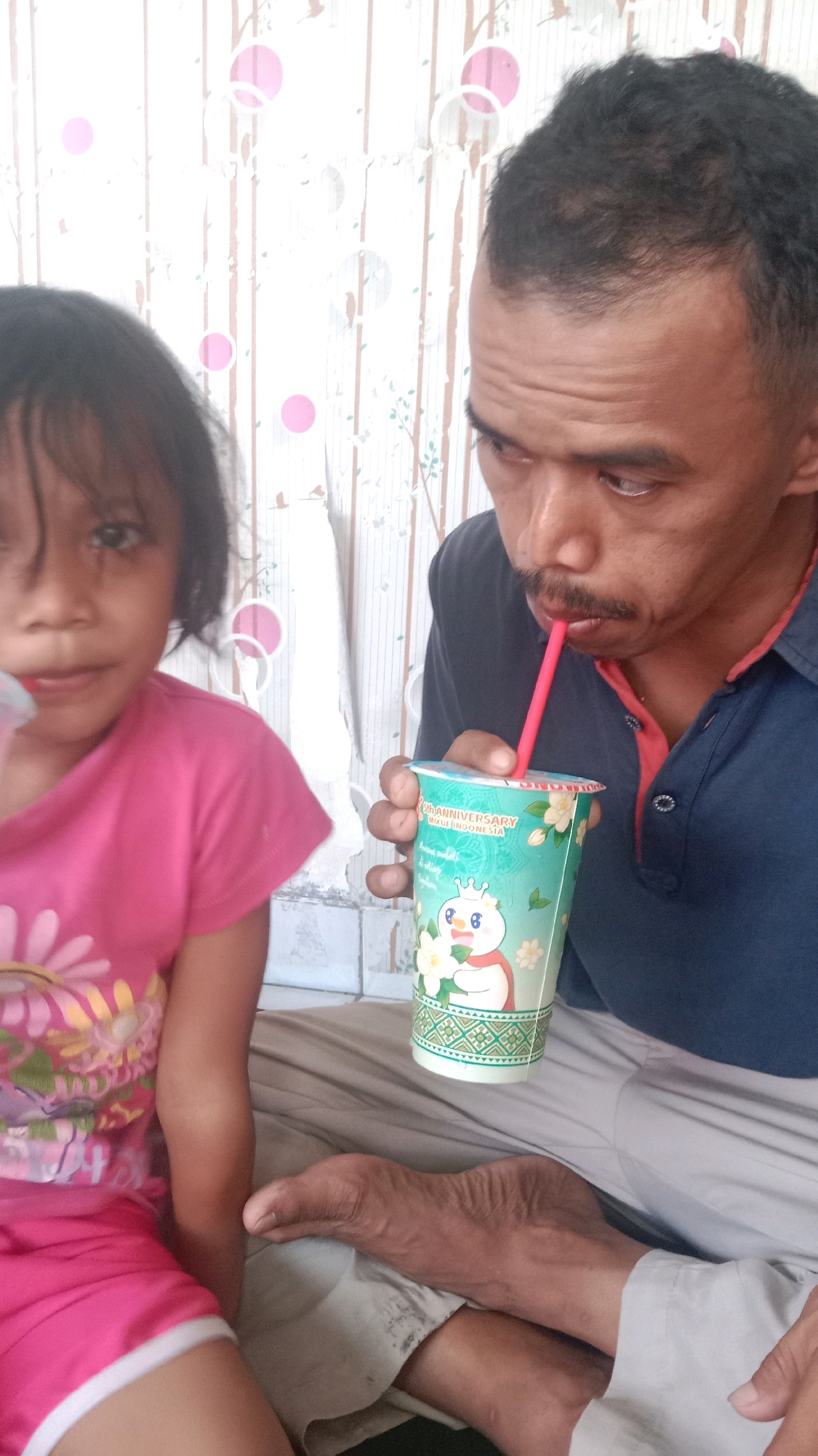 A man and a young girl are in a close-up portrait. The man is on the right, wearing a dark blue polo shirt with a red collar. He is holding a light green and white patterned cup with a red straw and drinking from it. The cup has an image of a snowman-like character and text that appears to say "20TH ANNIVERSARY" and "J.CO INDONESIA". His skin is dark, and he has short, dark hair with some receding hairline. His facial expression suggests concentration as he drinks.
To the left of the man, a young girl is partially visible. She has dark hair and is looking towards the camera with wide eyes. She is wearing a bright pink t-shirt with a large yellow flower design on it. Her left arm is visible, resting on her leg. Her skin tone is lighter than the man's.
They are positioned in front of a wall with a white, patterned wallpaper featuring vertical stripes and circular motifs in pink and light gray. To the right of the pair, a leg and foot of an adult, presumably the man, are visible, resting on light-colored pants. The environment suggests an indoor setting, possibly a home, in Sumberjaya, Indonesia. The lighting is even, and the focus is on the subjects and the cup.