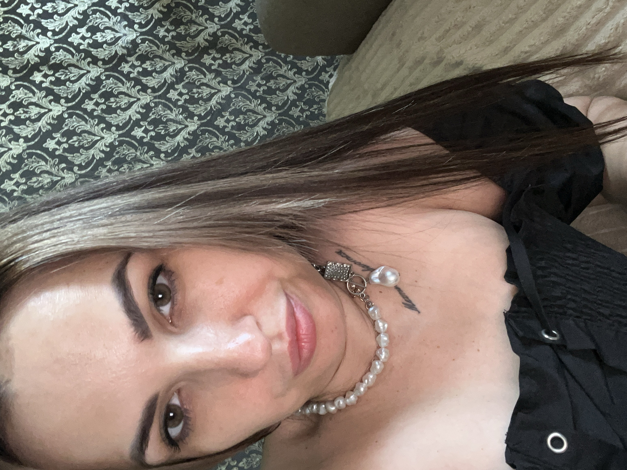 A woman with long brown and blonde hair is lying down. She is wearing a black top and a pearl necklace with a pendant. There is patterned fabric behind her and a light-colored cushion to her right. A tattoo is visible on her neck. The location is Unknown, Unknown.