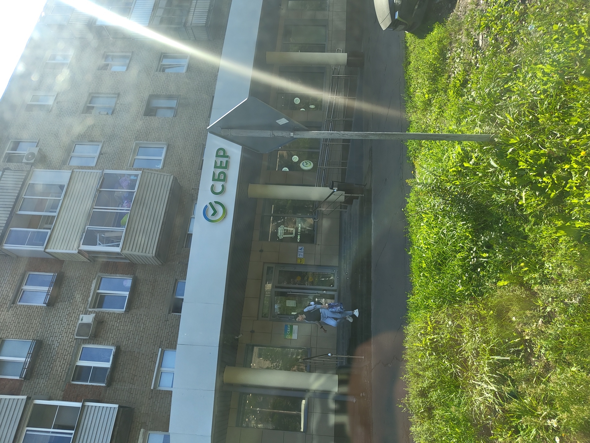 The image shows a Sberbank branch located in Zhukovsky, Russia. A woman wearing jeans and a dark top, carrying a large blue and white patterned bag, is walking out of the bank. She is on the steps leading out of the entrance, with her right foot forward. To the left of the entrance, a pillar supports the awning of the building. The bank's signage, featuring the Sberbank logo and text, is visible above the entrance. The building itself appears to be a multi-story residential building with cream-colored brickwork and multiple windows. In front of the building is a strip of green grass, and beyond that, a paved area, possibly a sidewalk or road. Sunlight is strongly illuminating the scene from the right, creating lens flare and reflections. A dark-colored car is partially visible on the right side of the image, parked on the paved area. A traffic sign on a metal pole stands in the foreground, slightly off-center.