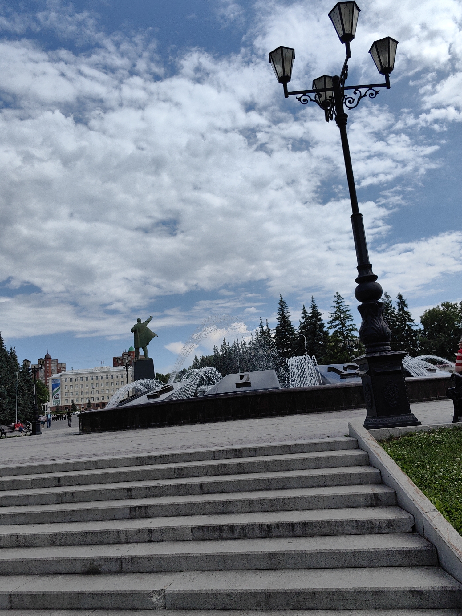 The image depicts a wide stone staircase leading upwards towards a large fountain with multiple jets of water spraying into the air. Beyond the fountain, a statue of Vladimir Lenin stands with his arm raised, pointing forward. In the background, a long, white building with many windows is visible, along with scattered trees and a cloudy blue sky. A lamppost with three lights is positioned prominently in the foreground on the right side of the image. A few people are walking and sitting in the distance, near the fountain and the building. This scene is located in the Lenin Square in Ufa, Russia.