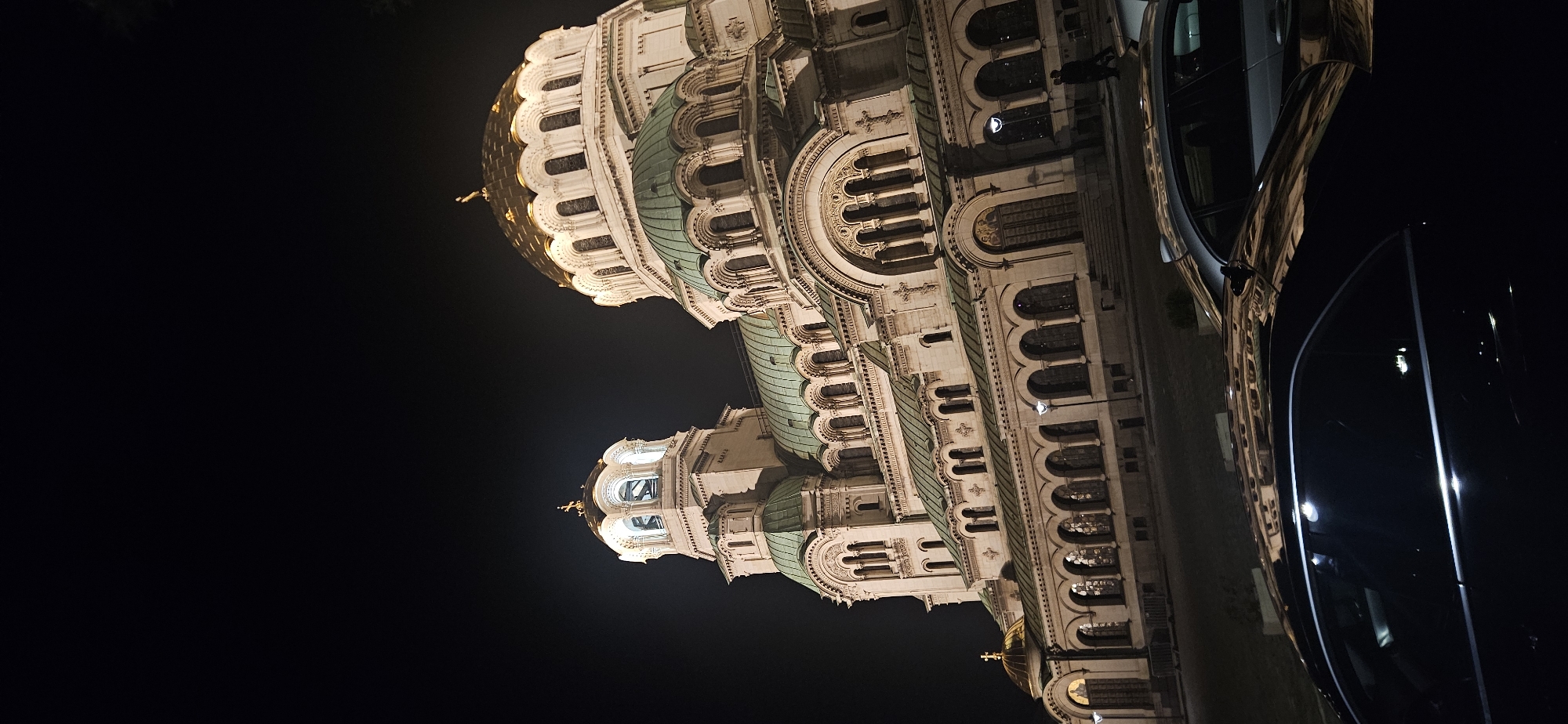 The image depicts the Alexander Nevsky Cathedral in Sofia, Bulgaria, illuminated at night. The cathedral's architectural details, including its domes, arches, and ornamentation, are visible. The sky is dark, contrasting with the illuminated building. Part of a vehicle is visible on the right side of the image. A person is standing next to the cathedral.