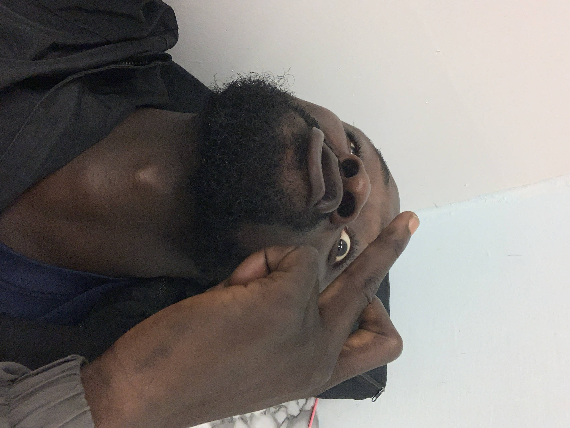 The image shows a dark-skinned man with a beard, looking upwards and to the right. His hand is raised, with his index finger extended. He is wearing a dark-colored jacket. The background is a plain white wall. There is no verifiable information to place the image in Lekki, Nigeria.