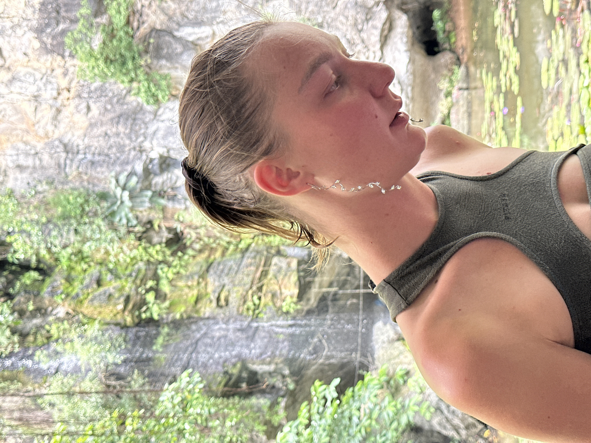 The media file shows a close-up, side-profile of a young person with their head tilted back and eyes closed, appearing to be in a state of relaxation or contemplation. They are wearing a dark green tank top and a delicate, silver-colored earring that drapes along their ear. The background is a blurred natural scene, suggestive of a lush, green environment with rock formations and water, possibly a waterfall or a pond with lily pads, consistent with a location like Nam Hoa Lư Ward, Vietnam. The lighting suggests it is daytime. The person's skin has a warm, flushed tone, and their hair is pulled back. The overall mood is serene and peaceful. There is no visible text in the image.