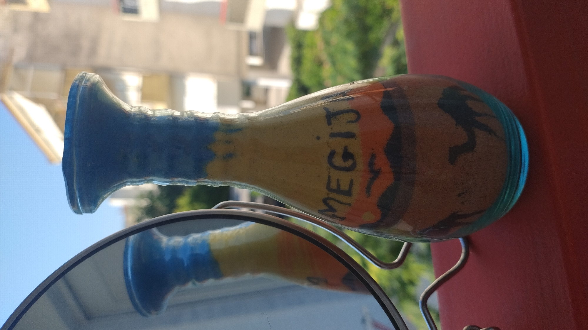 In the city of Perea, Greece, a decorative sand bottle with the inscription "MEGIJI" stands on a red surface, likely a balcony railing. The bottle is filled with colored sand layers, depicting a desert scene with camels and birds. A mirror reflects the bottle and its surroundings. In the background, buildings and greenery are visible under a clear blue sky.