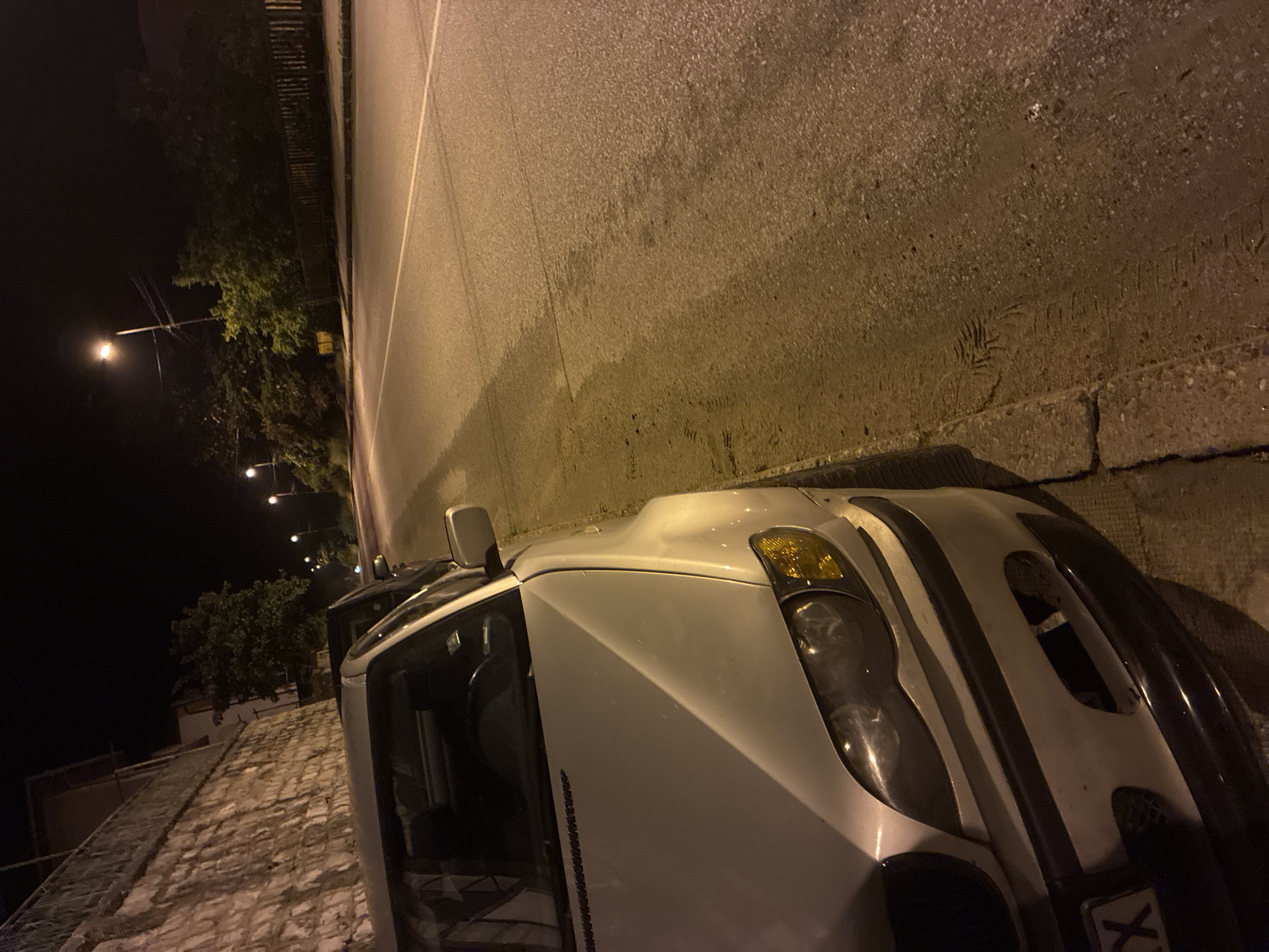 The image depicts a nighttime scene on a street in Vlahovo, Bulgaria. A light-colored, likely silver or white, BMW E46 sedan is prominently featured, parked parallel to the curb on the right side of the asphalt road. The front left quarter of the car is visible, showing clear headlights and an amber turn signal. The front bumper has black trim, and the lower grille opening on the passenger side of the bumper is missing. A partial Bulgarian license plate is visible on the lower left of the bumper, displaying "X" as the last character. The driver's side door appears to be open or ajar.
The asphalt road, illuminated by multiple streetlights casting a warm orange-yellow light, exhibits a single solid white line. The sidewalk adjacent to the car is paved with concrete. Distinct footprints and tire marks are visible in the dirt and dust along the edge of the sidewalk and on the road surface near the car.
To the left of the car, a tall stone retaining wall ascends, with a building and a metal balcony railing visible above it. In the background, dark trees line the street. Several streetlights illuminate the scene, casting shadows. Further down the road, another vehicle, darker in color, is partially visible behind the primary car. A wooden fence is also present on the right side of the road in the distance. No individuals or animals are visible in the scene.