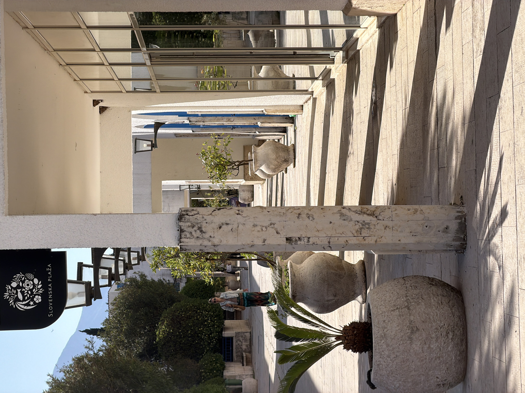 The image captures a sunlit corridor in Budva, Montenegro. The corridor is bordered by white, modernist architecture on one side, including a series of pillars and overhead glass panels, creating strong shadow lines on the tiled floor. On the other side, there are rows of potted plants, including trees and a palm, in large, aged stone pots. A sign marked "SLOVENSKA PLAŽA" is visible at the end of the corridor where vegetation and trees are present, suggesting a connection to a promenade or beach. A woman is visible in the frame.