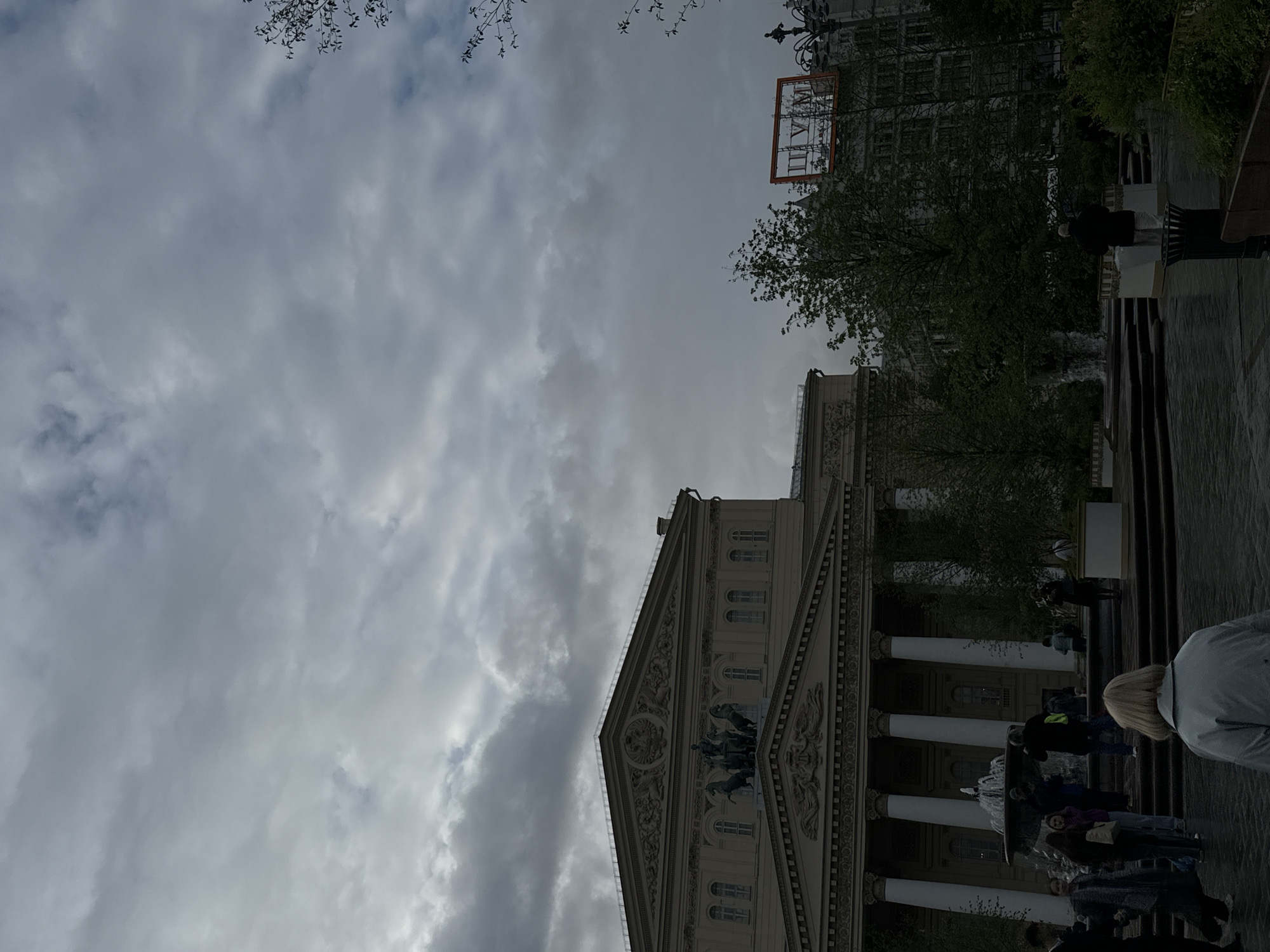 The image shows the Bolshoi Theatre in Moscow, Russia. The view is from a ground-level perspective, looking up at the building. The sky is overcast. A building with a sign is visible behind the theatre. People are present on the steps leading up to the building, and a fountain is near the base of the steps. The building is ornate with columns and sculptural details.