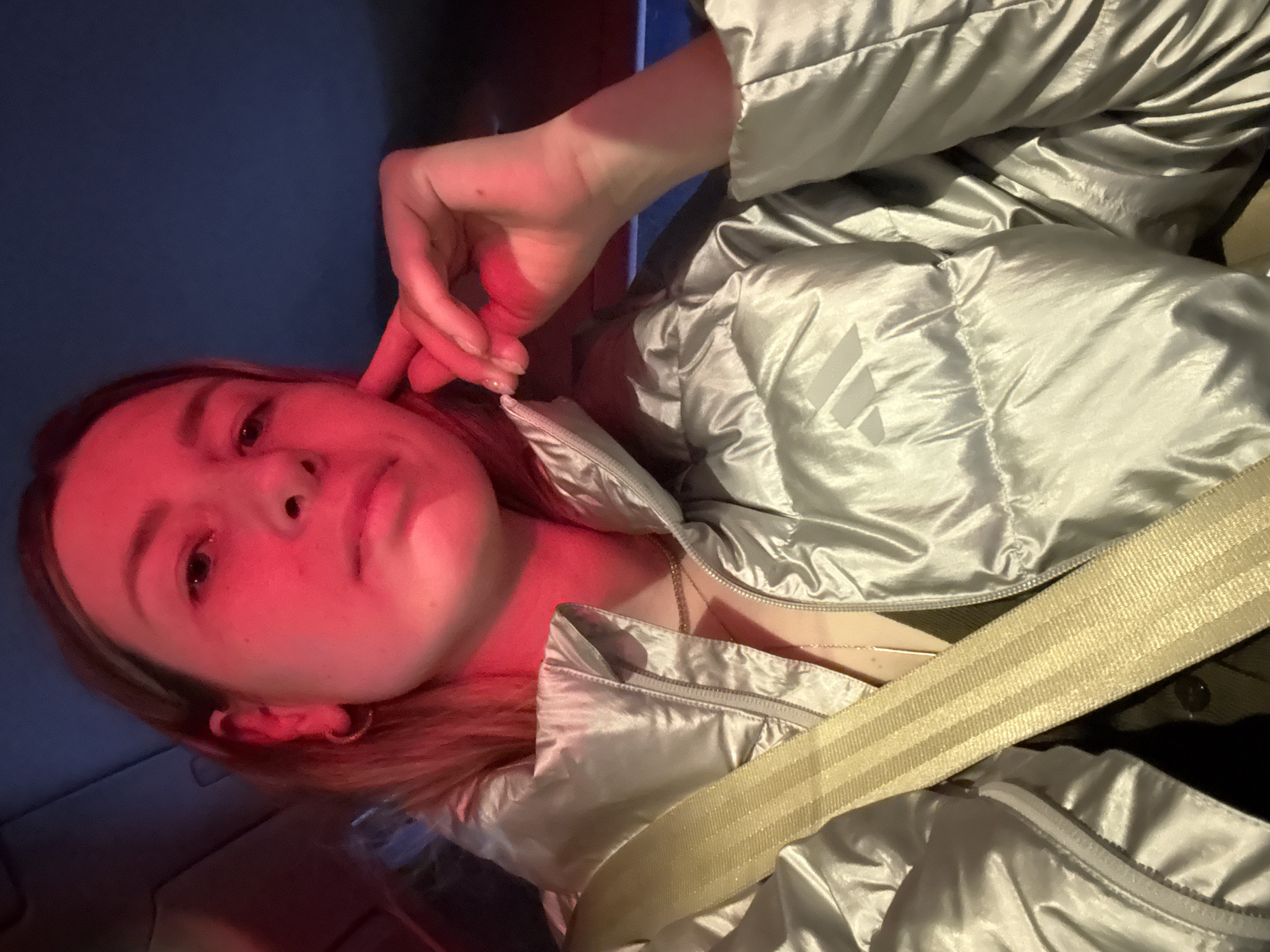 A young woman is in what appears to be a car, wearing a shiny silver jacket and a seatbelt. She is holding her hand near her face, and the lighting gives her skin a red hue. The background appears to be dark with a hint of blue. There is no information in the provided image to indicate that this picture was taken in the city of Moscow, Russia.