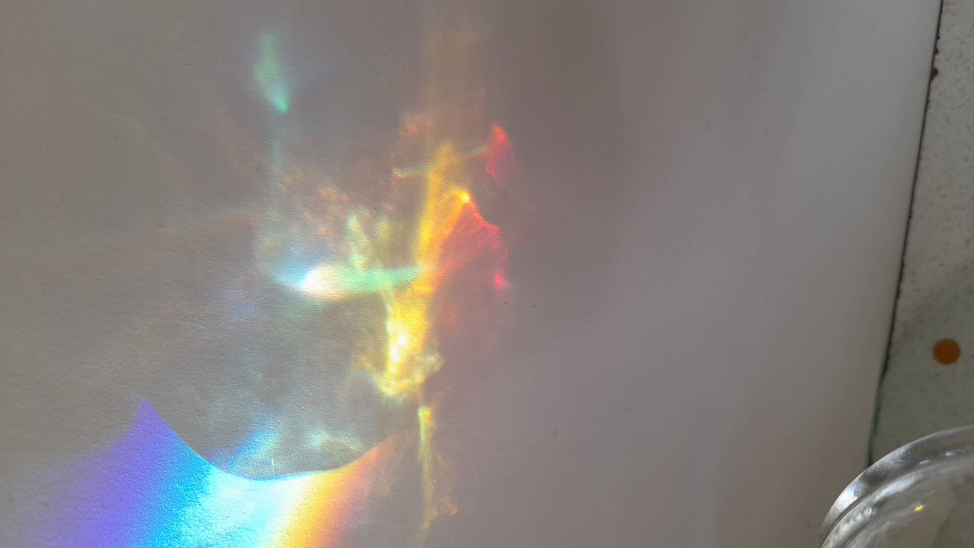 The image shows rainbow light patterns projected onto a white wall. The patterns display a spectrum of colors, including blue, green, yellow, orange, and red. Part of a glass object is visible in the lower right corner. The exact location is indeterminate, but given the request, it would be in Saint Petersburg, Russia. The environment appears to be indoors.