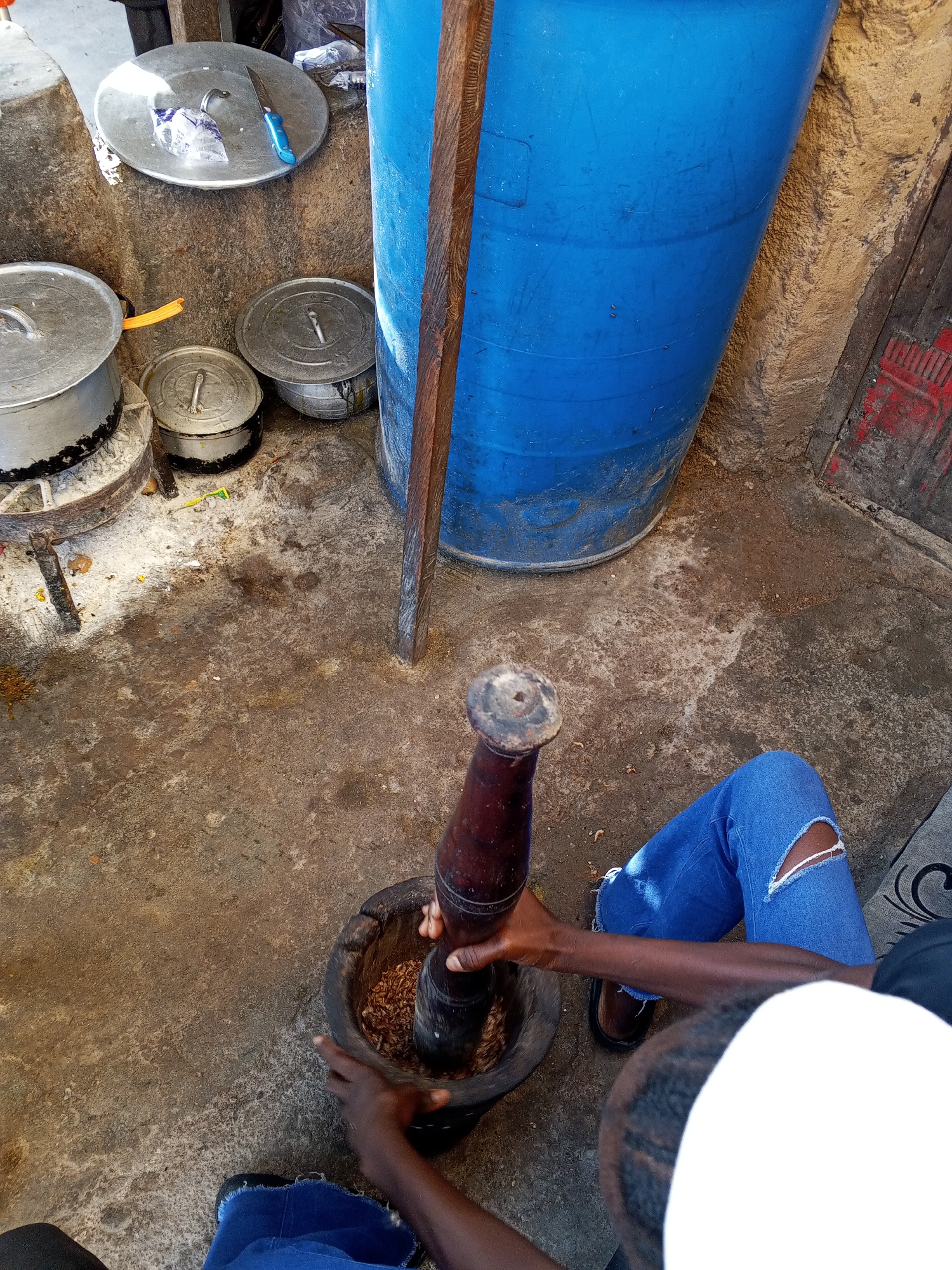 An overhead shot depicts a person in blue jeans with a rip in the knee, holding a wooden pestle and mortar. The person appears to be grinding or crushing items in the mortar. Near the mortar, there is a large blue plastic drum, various metal pots and lids, and a wooden beam. The scene takes place outdoors on a weathered concrete surface. Based on the prompt, the location is Gwong, Nigeria.