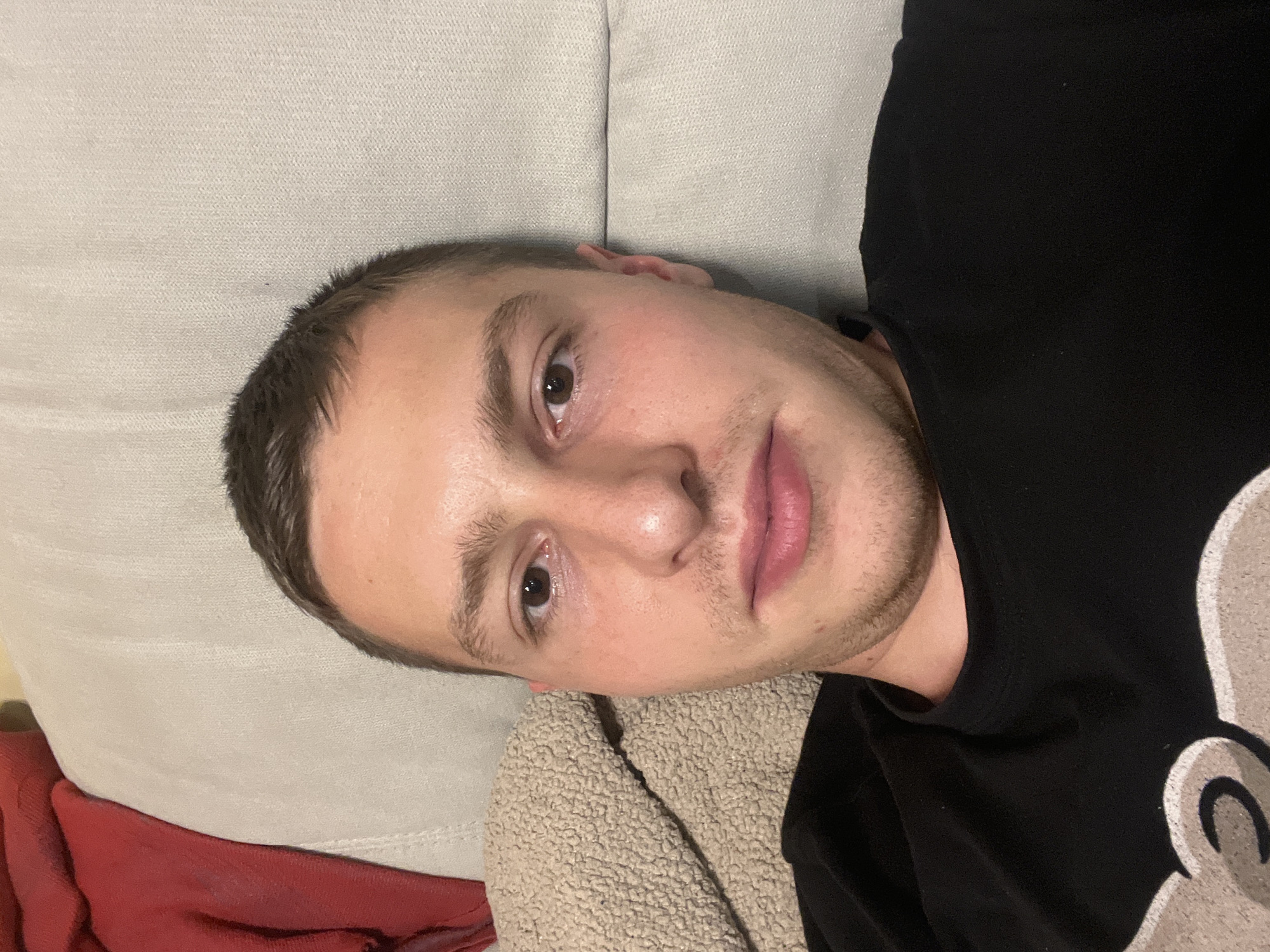 A young adult male with short dark hair and brown eyes is lying on a light gray upholstered surface, possibly a sofa. He is wearing a black t-shirt with a white graphic on the right side. His head is turned towards the camera. The background is blurred, but includes a red fabric item. The location of the photo is not discernible within the image itself, and Burgas, Bulgaria cannot be determined.