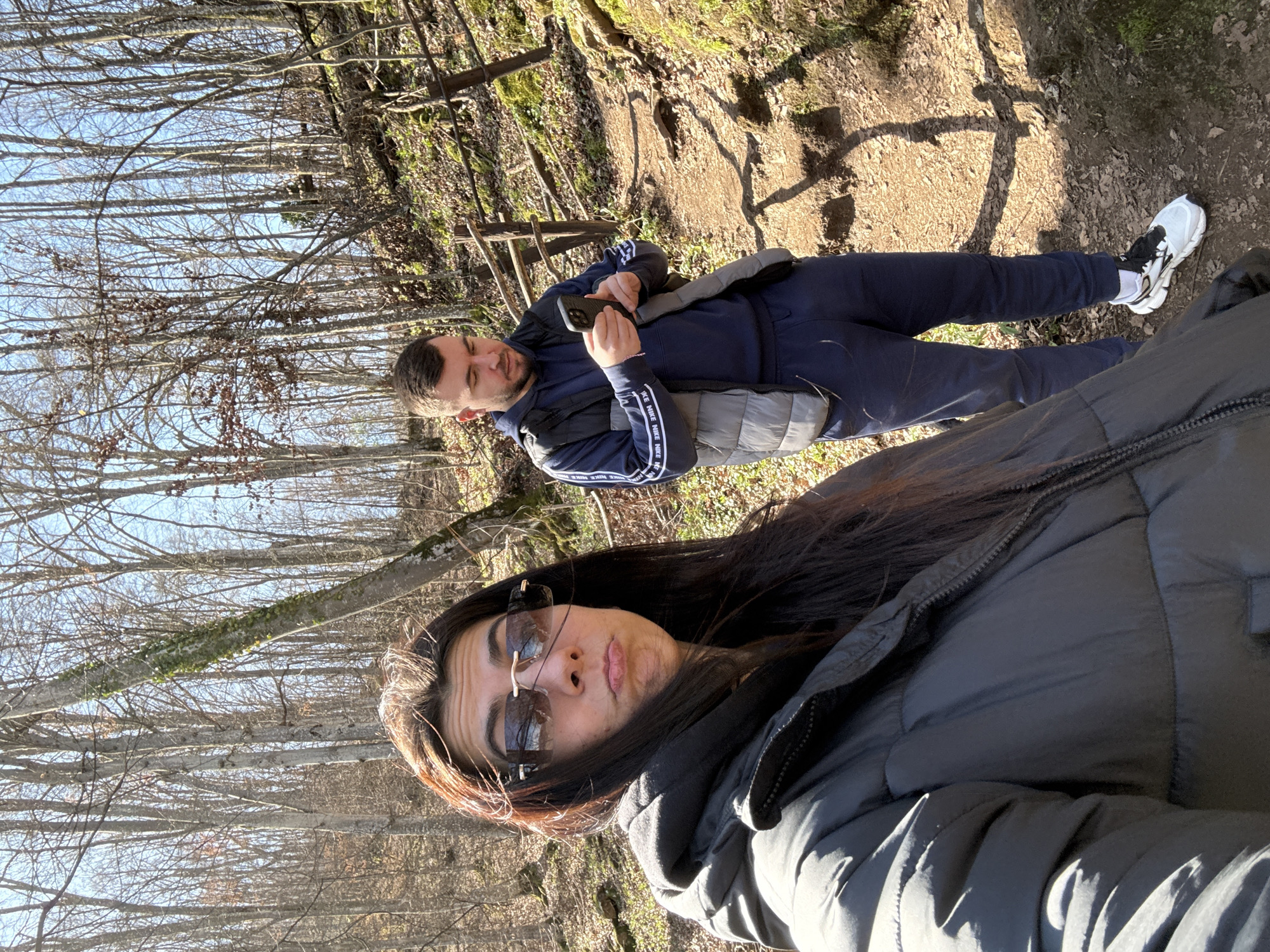 Two people are taking a selfie on a wooded path near Ribaritsa, Bulgaria. The person in the foreground is a woman wearing sunglasses and a gray puffer jacket. Behind her, a man is looking at his phone. He is wearing a blue tracksuit with white stripes and white sneakers. The path is dirt and leaves. Bare trees are visible in the background under a clear blue sky.