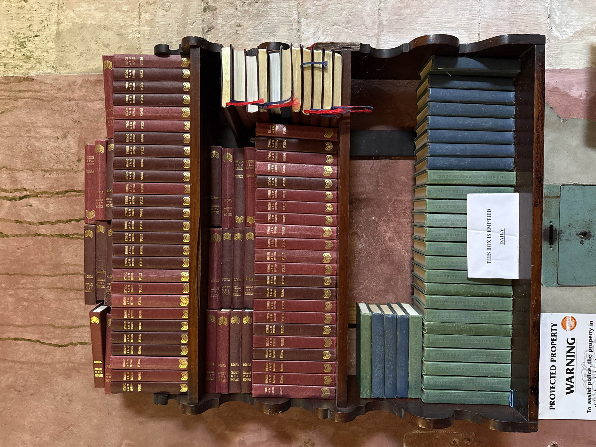 This image captures a weathered wooden bookshelf filled with numerous books, set against an aged, mottled wall. The scene is likely indoors, possibly within a church or historical building, given the nature of the books and warning sign.
The bookshelf, crafted from dark wood with decorative edges, holds books neatly organized into sections. The majority are dark red or maroon, featuring gilt lettering on their spines. These books are predominantly identified as "HYMNS A&M REVISED WORDS," suggesting they are hymnals from "Hymns Ancient & Modern." They are stacked vertically in multiple rows, with some additional copies laid horizontally on an inner shelf.
On the very top shelf, a smaller collection of lighter-colored books, possibly cream or light tan, are visible. Several of these books are adorned with red and blue ribbons, serving as bookmarks or ties.
To the right, a section of the bookshelf is dedicated to a stack of green and blue-covered books. These are identified as "THE SHORTER PRAYER BOOK," stacked vertically. Another small stack of similar books lies horizontally on a lower shelf to their left.
The background wall exhibits rough, textured surfaces with varying shades of beige, light brown, pink, and hints of green moss or discoloration, indicating age and possibly dampness. To the far right, a dull green metal panel with a keyhole is visible, resembling part of a historical fixture or a secure box.
Two white paper signs are affixed to the right side of the image. The upper sign clearly states: "THIS BOX IS EMPTIED DAILY." Below it, a partially visible warning sign reads: "PROTECTED PROPERTY WARNING SELECTADNA To assist police, the property in this church/area..." These signs, along with the nature of the books, strongly suggest a public religious setting in Great Yarmouth, United Kingdom, where these items are used and maintained.