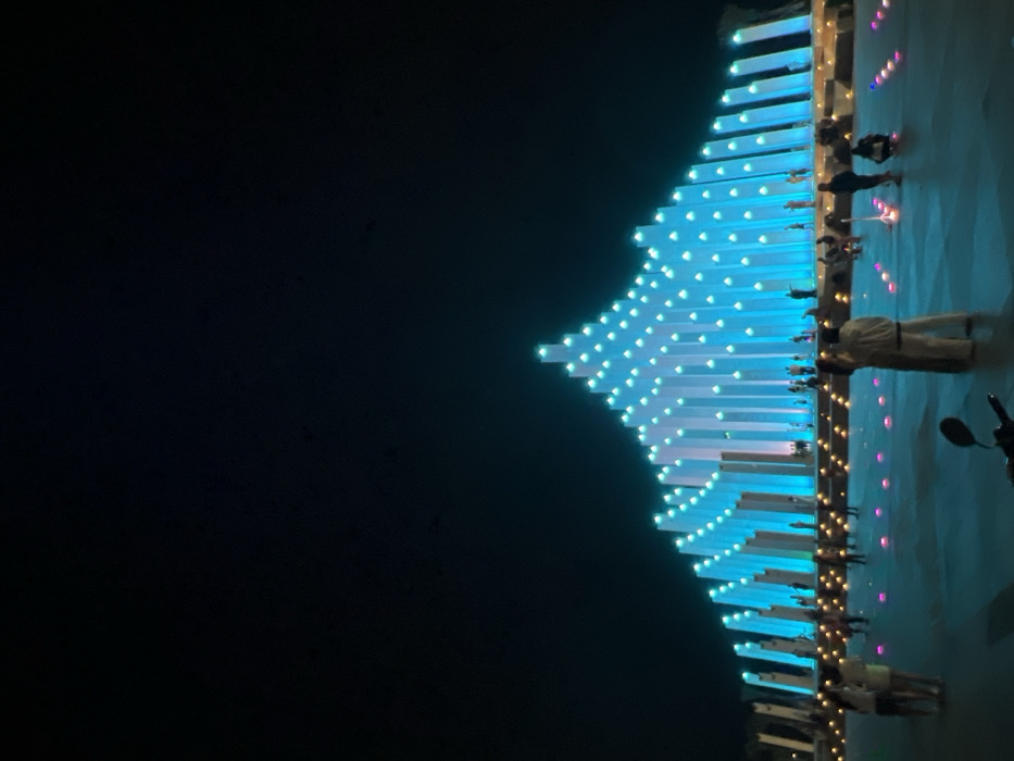 The image displays a nighttime scene within a public plaza in Ho Chi Minh City, Vietnam. The central feature is a large, architectural light installation composed of numerous vertical columns of varying heights, arranged to form a central peak that gradually descends towards both sides. These columns are illuminated with vibrant blue and white light, with individual light sources visible on their upper surfaces. The structure is silhouetted against a dark, unlit sky.
The ground area of the plaza is paved and diffusely lit with a blue hue, additionally featuring numerous scattered, spherical ground lights in yellow, pink, and blue tones. Approximately 30 to 50 individuals, appearing to be adults and some children, are present throughout the plaza. They are observed walking, standing, and gathering in small groups, primarily facing or observing the illuminated structure. In the lower-left foreground, the black front fender and handlebars of a motorcycle are partially visible.