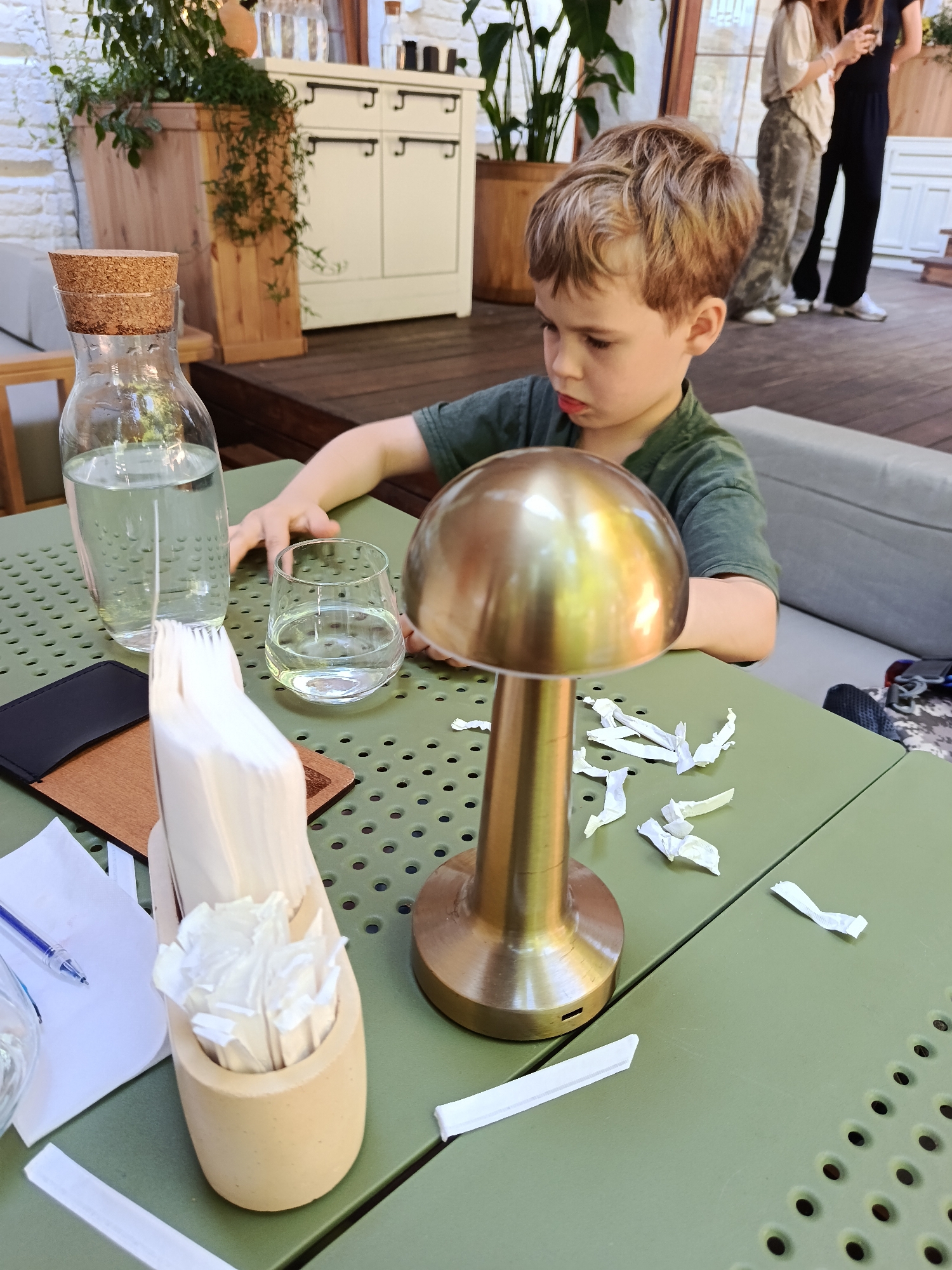 In Kyiv, Ukraine, a young boy sits at a green, perforated outdoor table. He wears a green t-shirt and has light brown hair. In front of him, a brass mushroom-shaped lamp stands on the table, surrounded by crumpled white paper wrappers. To his left, a glass carafe filled with water has a cork stopper. Next to the carafe is a glass of water. A wooden holder contains several white paper packets, likely for sugar or napkins. A black wallet and a pen are also visible on the table. In the background, two women are standing, one in a black outfit and the other in patterned pants and a light-colored top. A white cabinet with black handles is situated behind them, and lush green plants are visible. The setting appears to be an outdoor seating area of a cafe or restaurant.