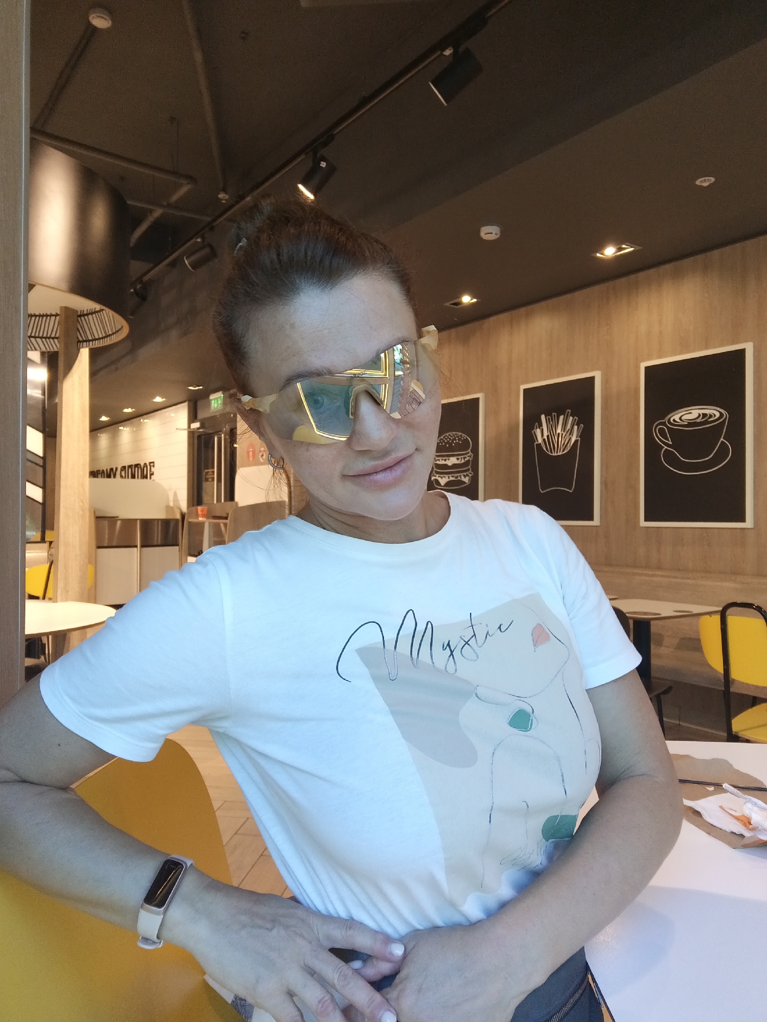 A woman is pictured indoors, likely within a fast-food restaurant environment, given the wall decor featuring illustrations of a burger, french fries, and a cup of coffee, and the presence of bright yellow seating. The woman is wearing a white t-shirt with a graphic print that includes the word "Mystic" and an abstract line drawing of a face. She also has on large, tinted sunglasses. Her hair is pulled back in a bun. In the background, there are tables and chairs, along with lighting fixtures on the ceiling. The overall lighting suggests an indoor setting. The context of Sochi, Russia, is not visually present within the image itself.