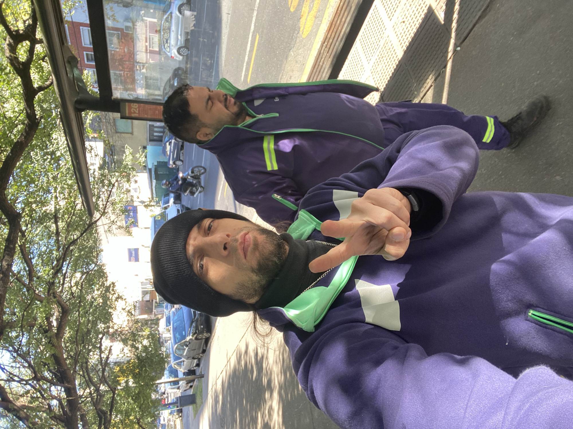 Two men wearing purple tracksuits are posing in a public area. One man is standing and gesturing towards the camera while wearing a black beanie. The second man is lying down next to him. They are located near a bus stop, evidenced by the metal structure and bus schedule signage. Cars and trees line the street. The location is Buenos Aires, Argentina, based on the street signage.