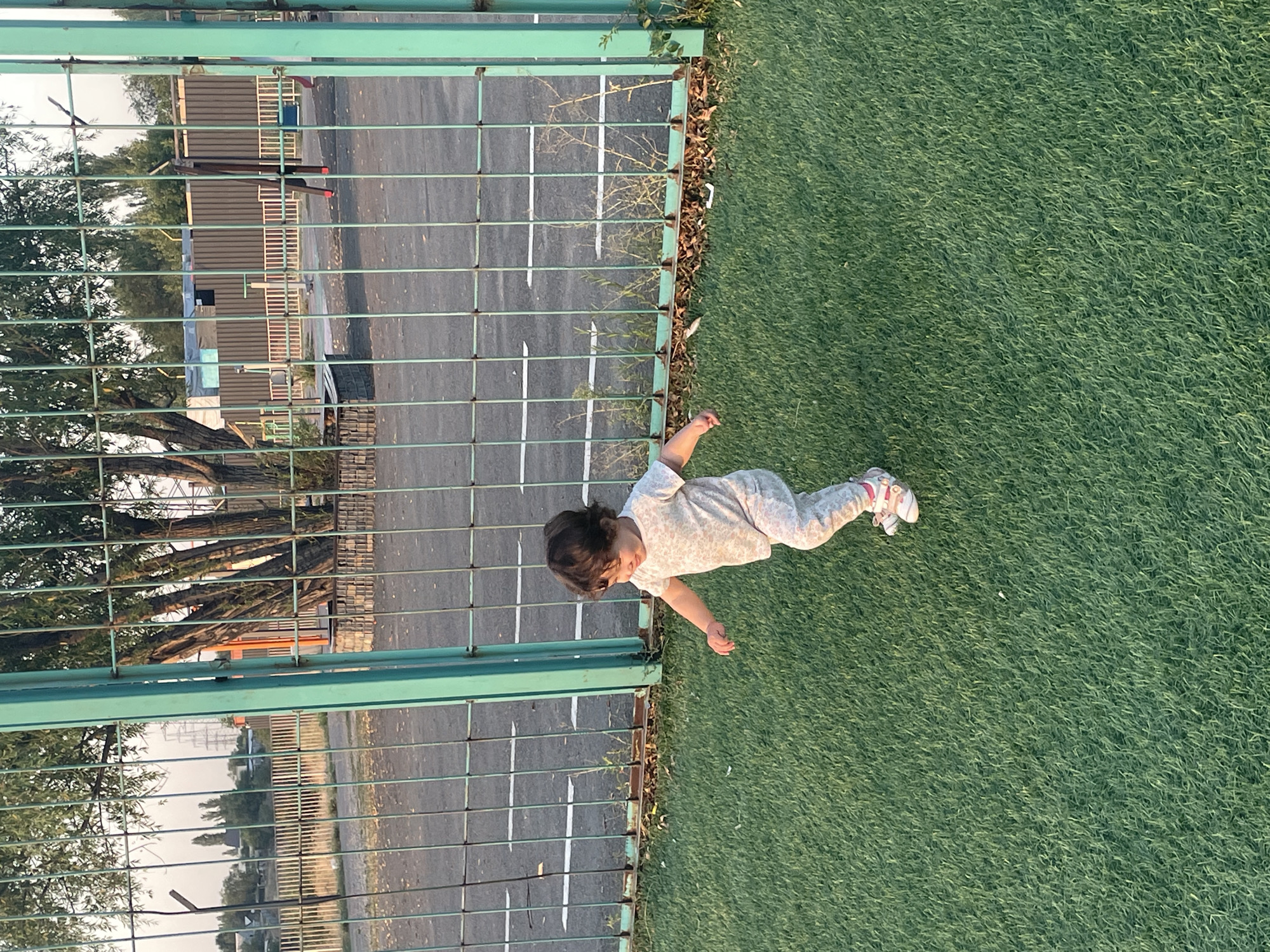 A young child, likely a toddler, is captured mid-stride on a vibrant green artificial turf surface. The child is wearing a light-colored, patterned onesie and pink and white sneakers. They appear to be walking or running towards the right side of the frame, with their head turned to the left. Behind the child, a metal fence with green posts separates the turf area from a paved surface marked with white lines, suggesting a road or sports court. Beyond the paved area, trees with dense foliage are visible, and further in the distance, buildings and electrical infrastructure can be discerned against a hazy sky. The scene suggests an outdoor recreational area, possibly a park or playground, within a semi-urban or industrial setting. The light suggests it is either early morning or late afternoon. The context of Almaty, Kazakhstan, places this scene in a large city known for its mountainous backdrop and a mix of modern and older architecture, which aligns with the visual elements present.