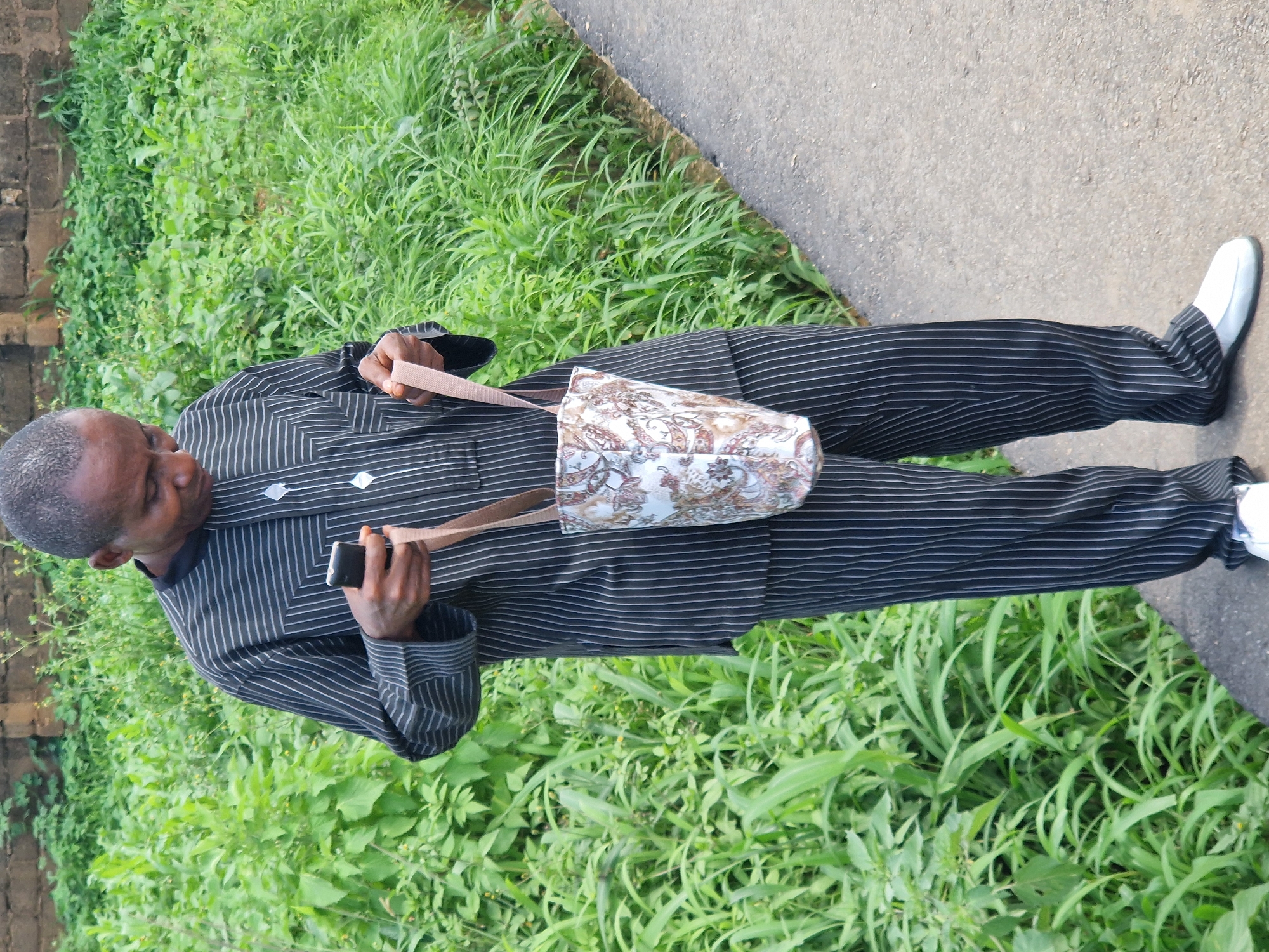 A man wearing a black and white pinstripe traditional outfit stands outdoors on an asphalt path next to lush green foliage. He is holding a smartphone in his right hand and a patterned bag with a light brown strap in his left. The man is looking down at his phone. The background consists of a brick wall and dense vegetation. The lighting suggests it is daytime.