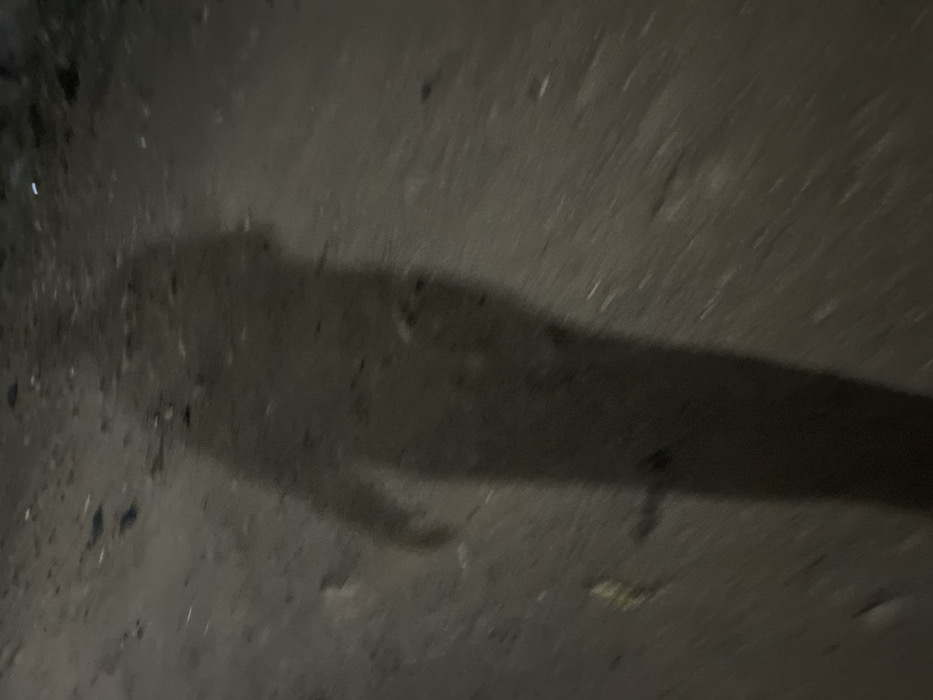A shadow stretches across a rough, dark gray surface, possibly asphalt or concrete. The shadow's shape is elongated, with a vague outline resembling a figure or object casting it. Irregular spots and patches of varying darkness are scattered across the surface, adding texture and depth to the scene. The lighting is dim, casting the entire composition in a subdued and somewhat mysterious atmosphere.