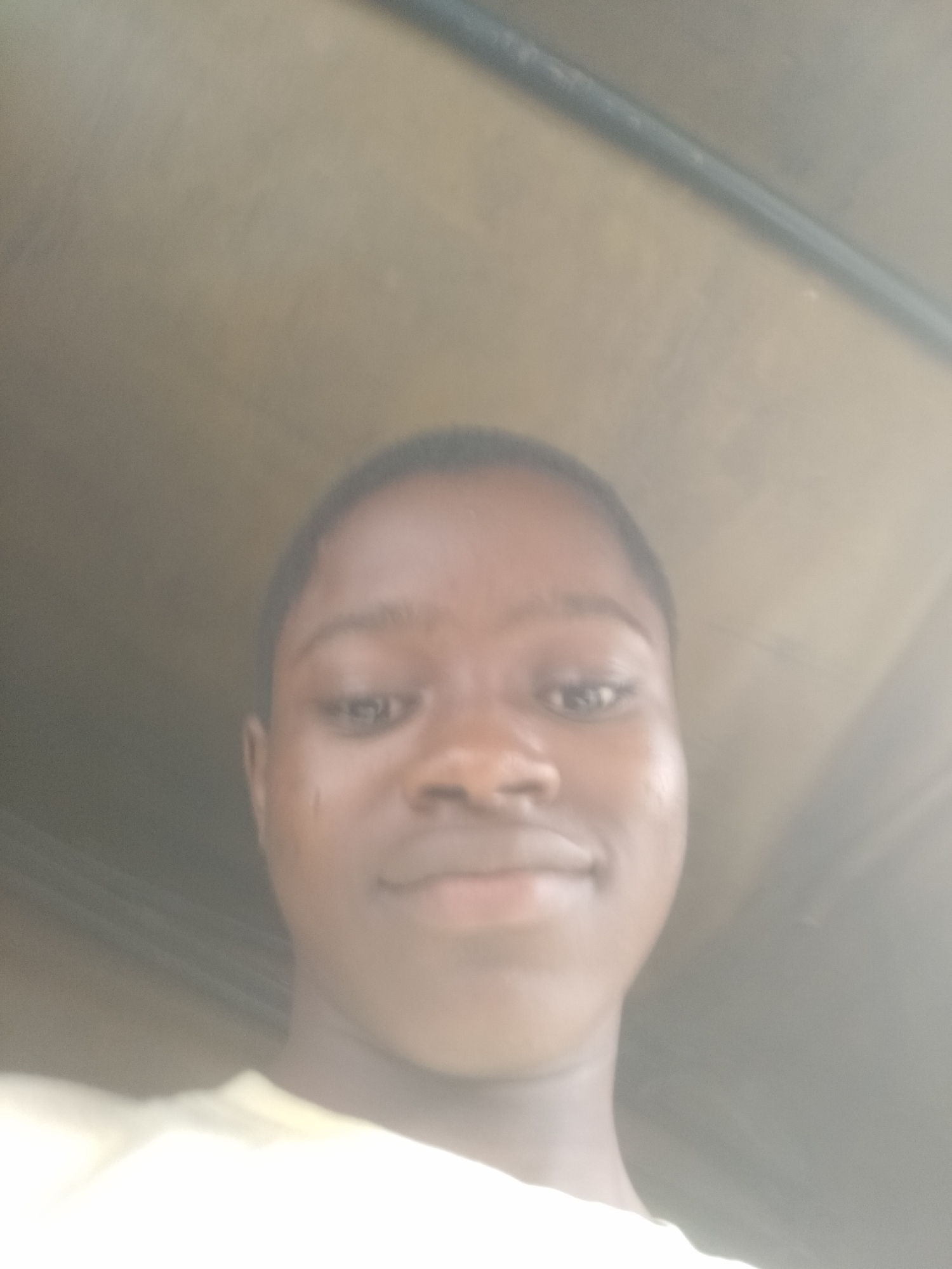 A medium close-up shot captures a person with dark skin and short, dark hair. The person is positioned in the lower center of the frame, their face angled slightly downwards towards the camera. They appear to be wearing a light-colored shirt. The background is out of focus and features what seems to be a gray or tan-colored wall or ceiling, with some darker elements suggesting a doorway or frame above. The location is assumed to be Shangisha, Nigeria, based on the prompt's parameters.
