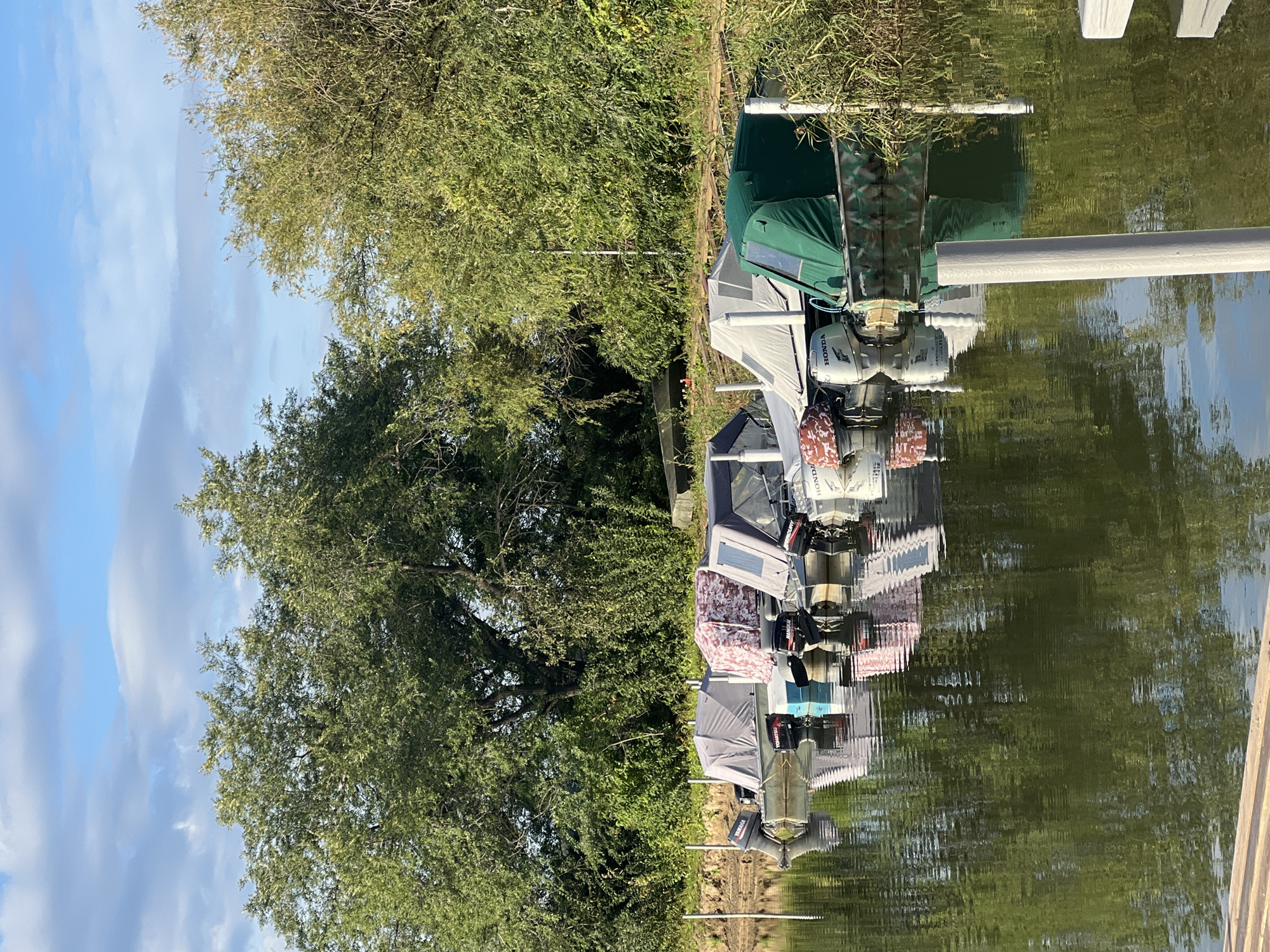 The image depicts a serene waterfront scene, likely in the vicinity of Novopichugovo, Russia. A line of boats is moored along the shore, their outboard motors visible. Each boat is covered with a tarp, some of which have patterned designs. The water reflects the boats and the lush green trees that form a dense backdrop. The sky above is partly cloudy, with patches of blue peeking through. The overall impression is one of tranquility and stillness, suggesting a quiet morning or evening by the water.