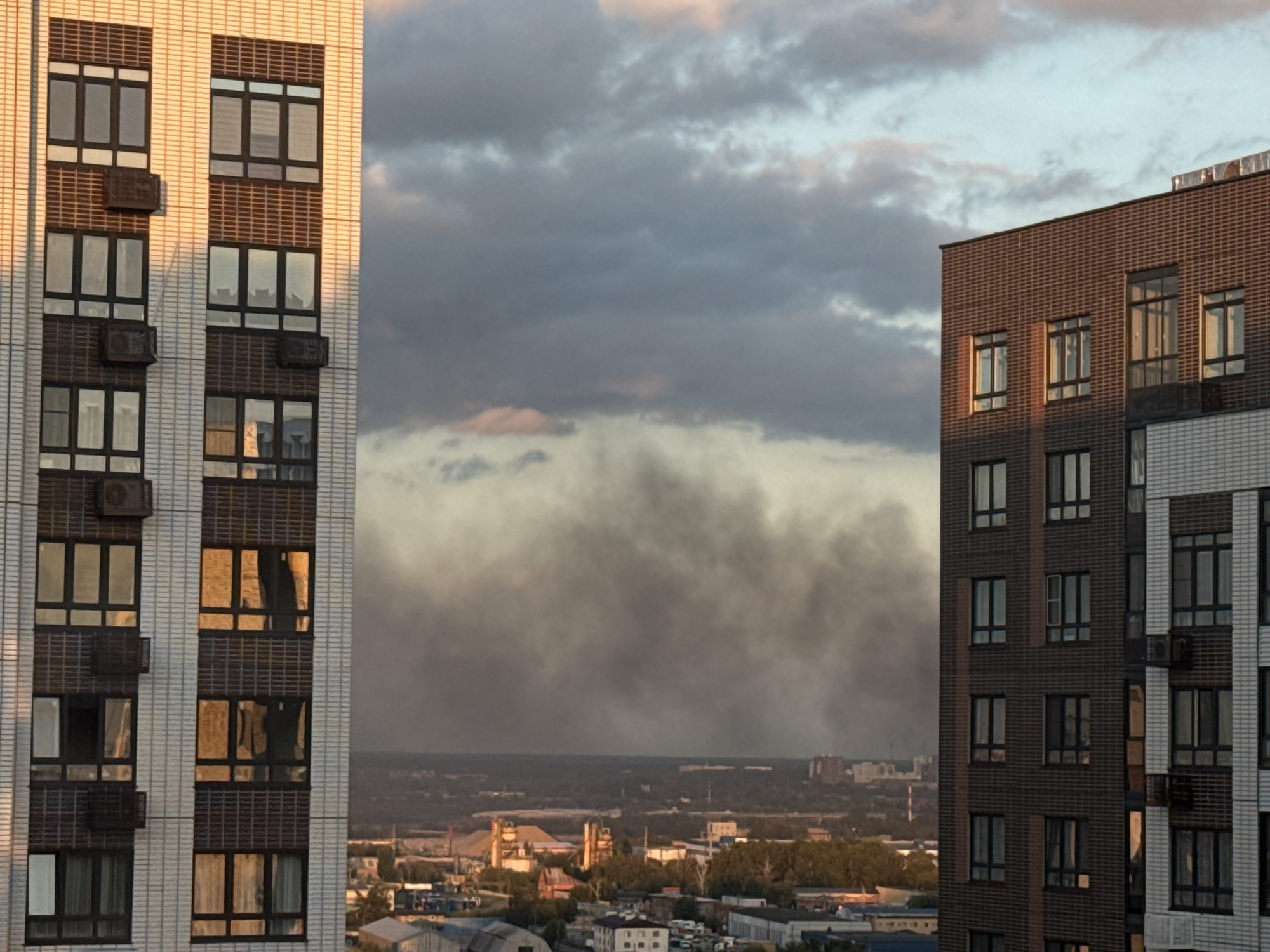 In Lyubertsy, Russia, an aerial view captures a smoky plume rising from industrial structures and dispersing into the sky. The scene is framed by the facades of two modern apartment buildings, one light-colored and the other dark brick, positioned in the foreground. The architecture features numerous windows and external air conditioning units. The background reveals a dense urban landscape with lower-rise buildings and interspersed trees, all under a dramatic sky filled with heavy, dark clouds tinged with pink and orange from the setting sun.