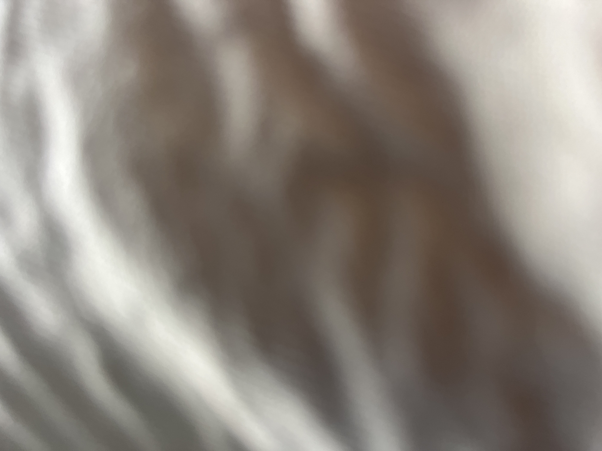 The image presents a highly blurred, abstract composition. The scene, located in the city of Casablanca, Morocco, depicts an undulating surface characterized by soft transitions between light and shadow. Dominant hues include white, various shades of grey, and muted brown, indicative of a soft, folded material or surface. Bright highlights are prominent in the upper-middle and right, contrasting with darker, warmer tones in the lower and left portions. The lighting appears diffused, creating smooth gradients across the surface. No discernible people, specific objects, actions, or interactions are identifiable within this close-up view.
