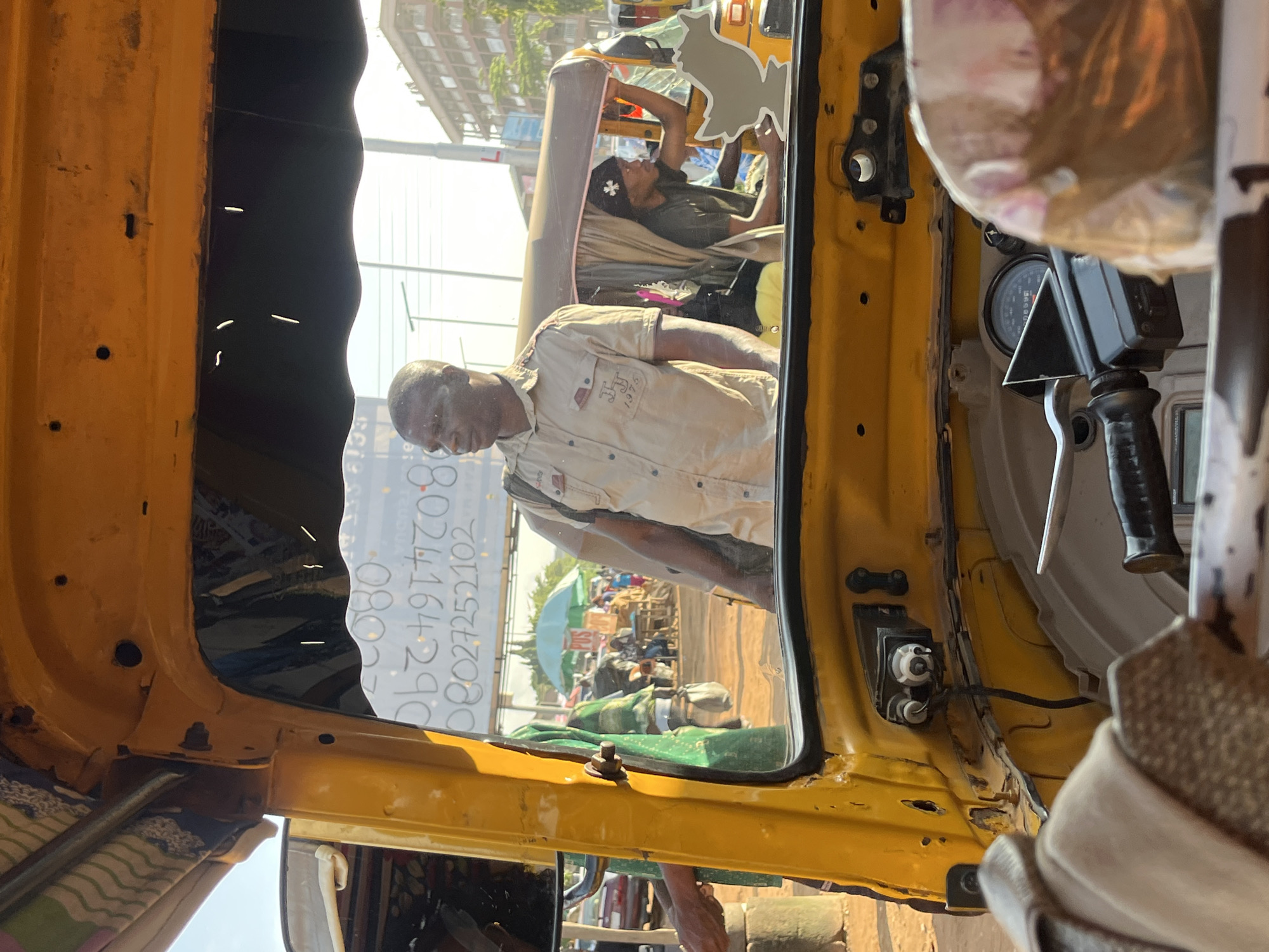 A man in a light-colored shirt stands near the open door of a yellow auto-rickshaw, his left arm visible. Behind him, another auto-rickshaw is partially visible with a person in a dark shirt and black beanie. The scene appears to be outdoors with a clear sky and distant buildings suggesting an urban environment, possibly in Abuja, Nigeria. The auto-rickshaw's interior shows a steering column and dashboard. In the background, a green umbrella and several people are visible, along with what appears to be a market stall. The overall lighting suggests it is daytime.