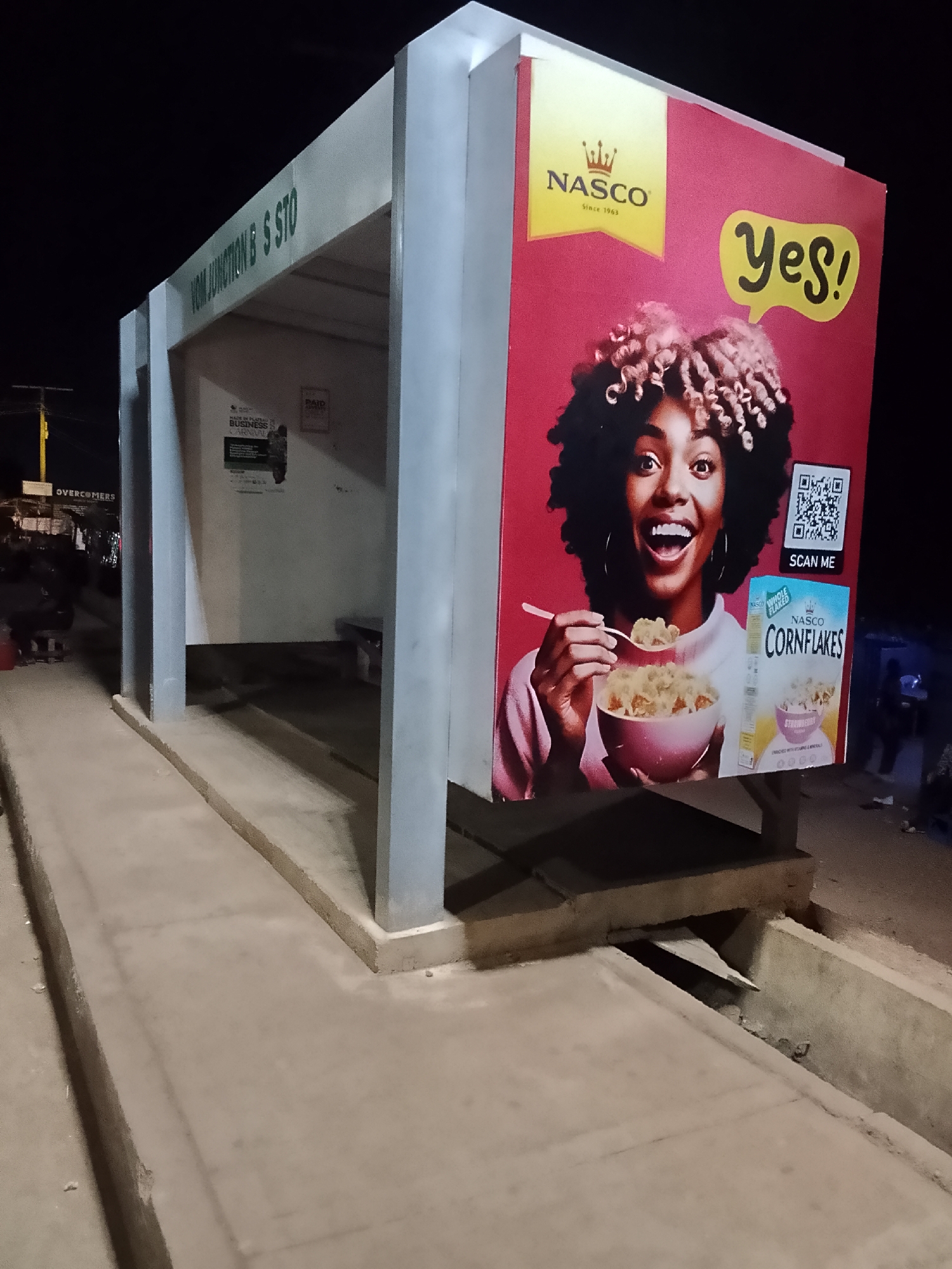 The image shows a bus stop in Bukuru, Nigeria, at night. The bus stop is constructed of gray concrete and has a large advertisement on one side for Nasco Cornflakes. The advertisement features an image of a smiling woman eating cornflakes and a QR code that says "Scan Me." The words "Vom Junction B S Sto" are painted on the front of the bus stop. In the background, there are people and vehicles. The location is Vom Junction Bus Stop in Bukuru, Nigeria.