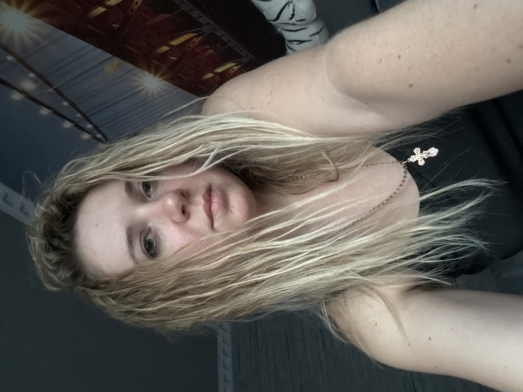 An indoor, close-up selfie of a young blonde woman with wet hair, wearing a black tank top and a silver cross necklace. The background includes a dark wall on the left and a mural of a bridge with lights on the right. The woman is looking slightly off-camera.