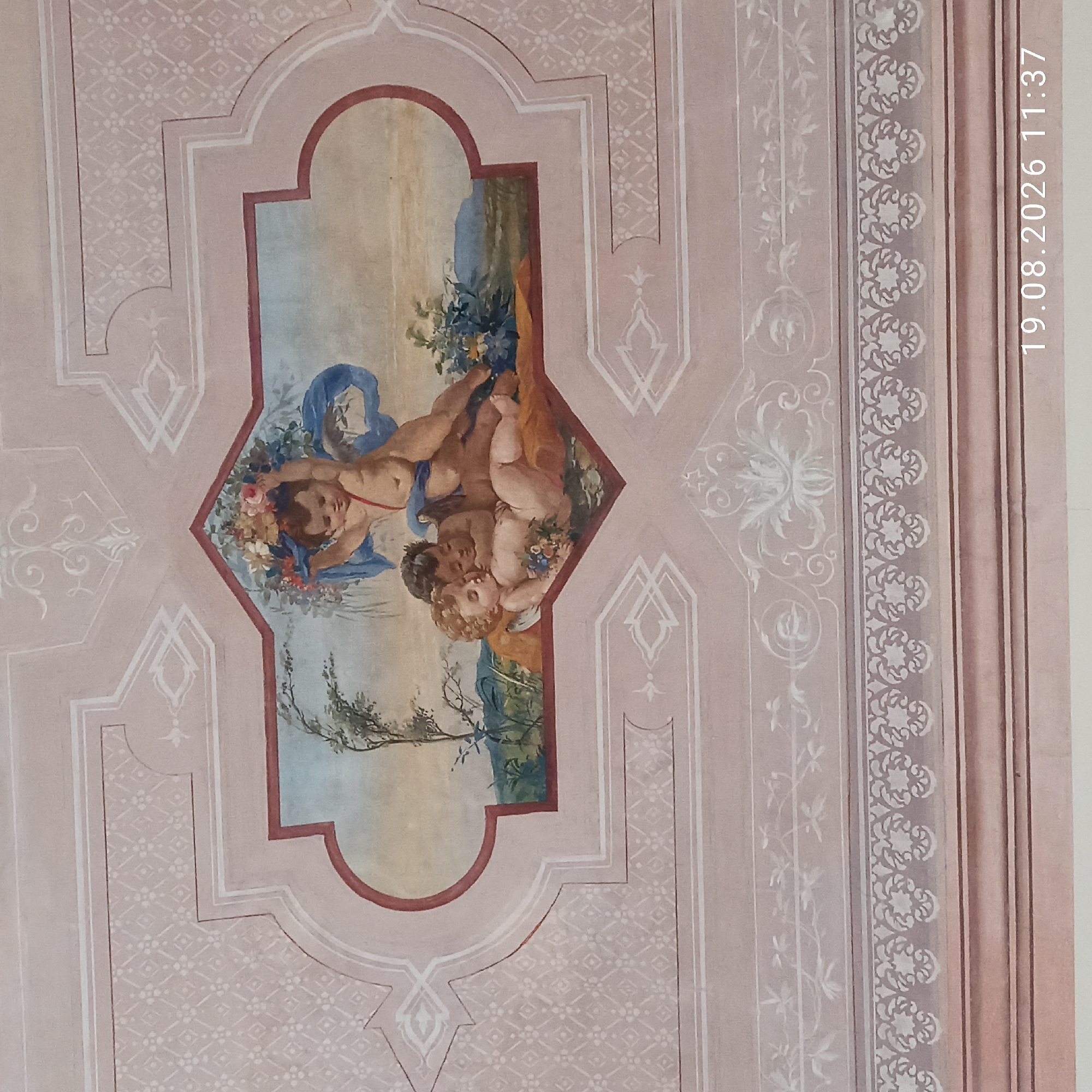 The image shows a fresco on a ceiling depicting three cherubs in a landscape setting. The cherubs are light-skinned, plump, and have curly hair. One cherub is at the top, holding a bouquet of flowers and a blue fabric. Below, two cherubs are nestled together, one lying on its back and the other leaning on it. The one on its back holds a small bouquet of flowers. The background of the fresco includes a tree, foliage, hills, and a pale sky. The fresco is framed by decorative borders with geometric and floral motifs in shades of pink, white, and reddish-brown. The surrounding ceiling also features ornate white patterns. The bottom right corner of the image shows a timestamp "19.08.2026 11:37". This fresco is located in the Sala degli Affreschi (Room of Frescoes) in the Palazzo Caputi in Ruvo di Puglia, Italy.