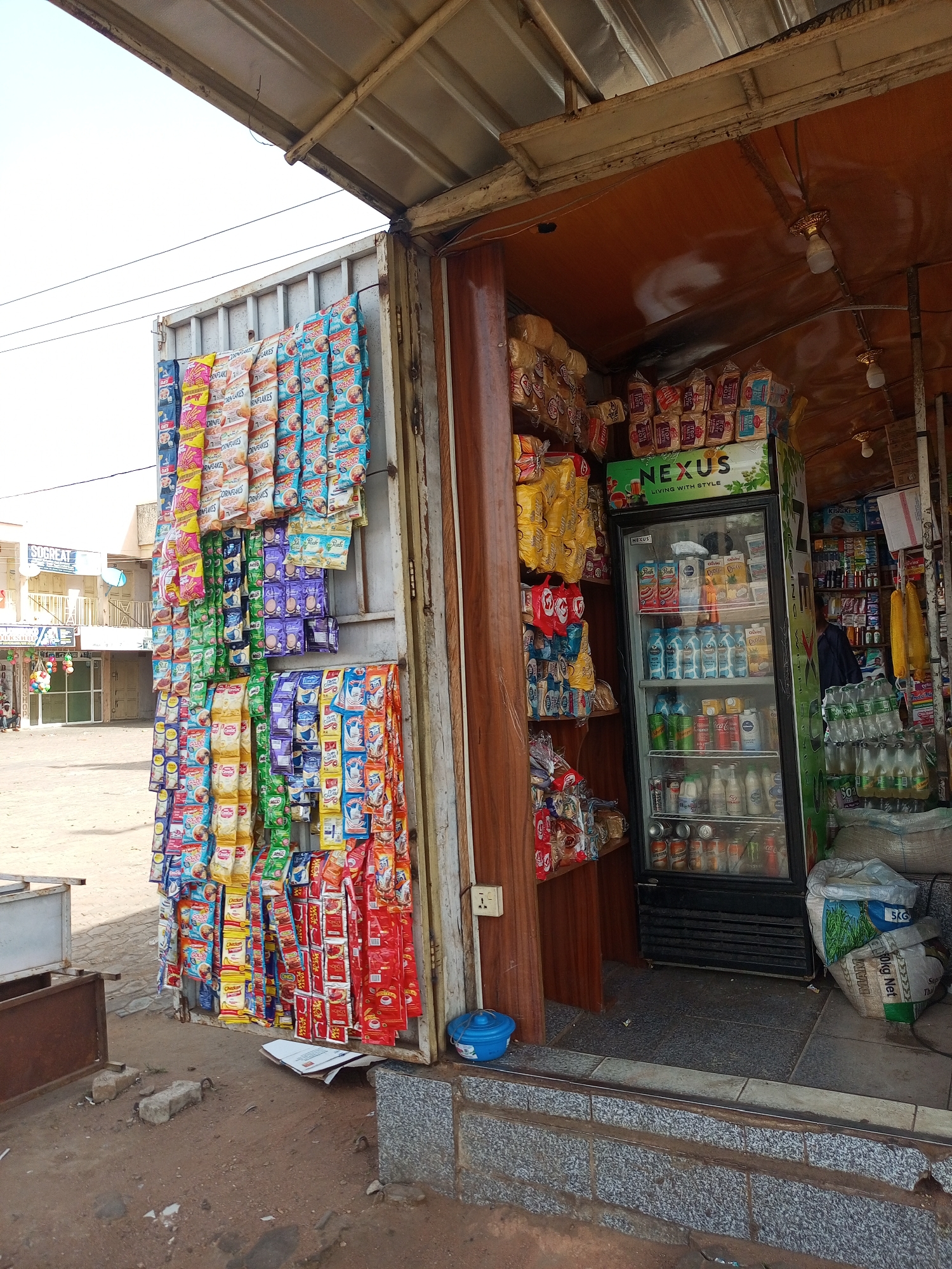 A well-stocked retail kiosk in Gombe, Nigeria, displays a variety of goods. Packets of instant products like "Cornflakes" and "Peak" milk are suspended from the exterior metal frame, showcasing the store's merchandise to passersby. A "Nexus" branded refrigerator, filled with chilled beverages, is positioned at the entrance. Shelves inside are stocked with bread, milk and other household essentials. Sacks of commodities are stacked near the entrance. The kiosk is open to the elements, with a simple step leading up to the interior. The surrounding area includes a street and the facade of other buildings.