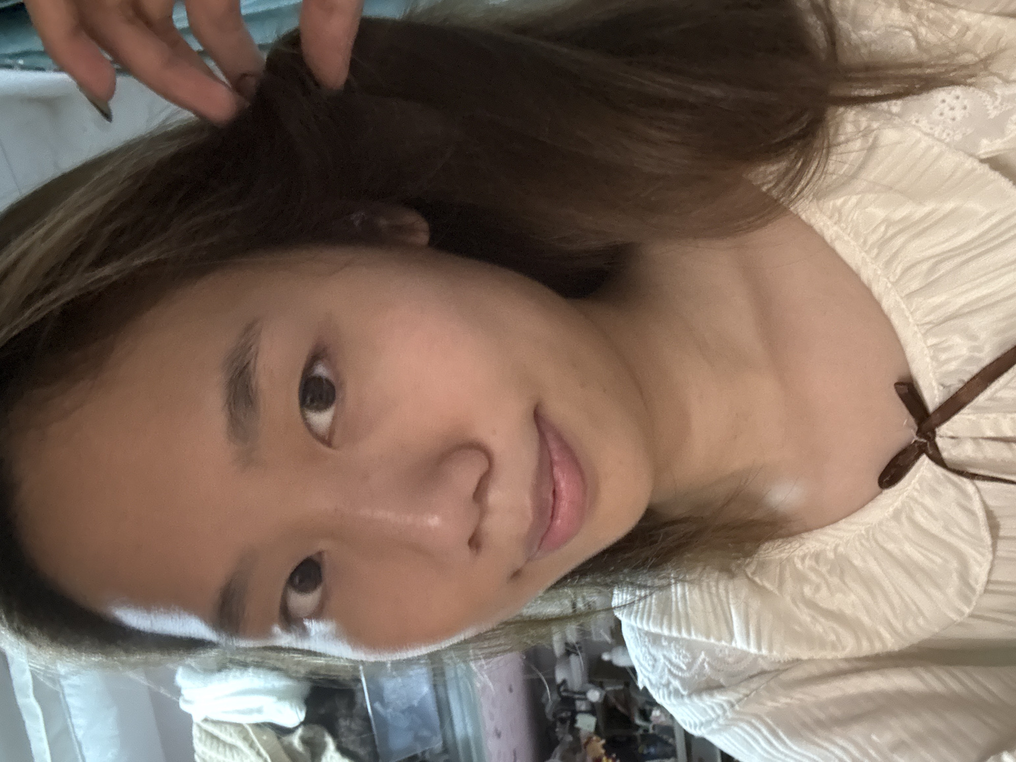A medium close-up shot shows a person with brown hair and light skin, likely female, looking at the camera. A hand with dark fingernails is touching their hair. They are wearing a white, ruffled top with a brown ribbon. The background is blurry, suggesting an indoor environment. There is no information in the image to indicate the location is in Nan'an District, China.