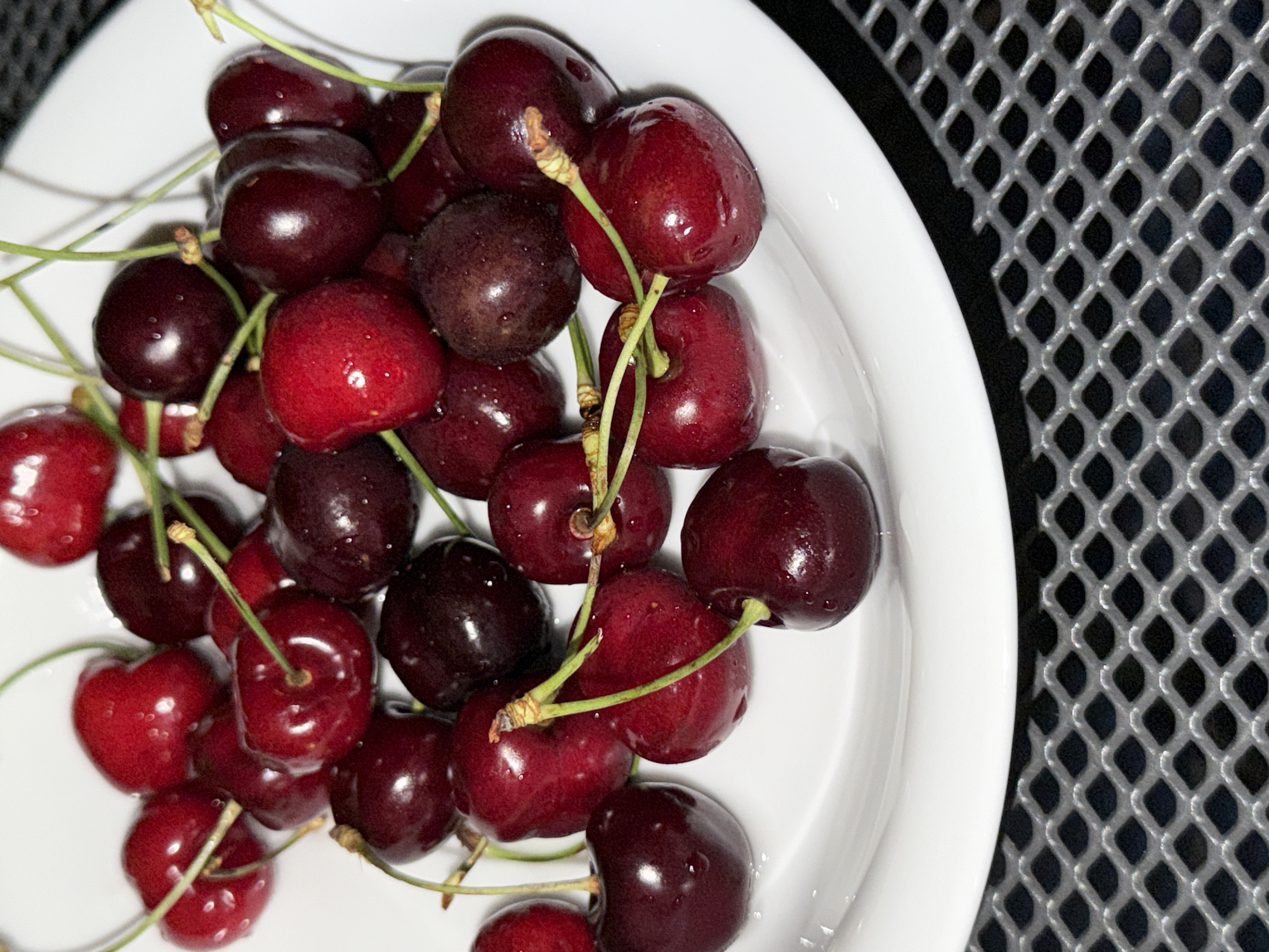 The image provides a close-up view of a substantial quantity of fresh cherries resting in a plain white, round dish. The cherries display a range of colors, from vibrant red to deep, dark maroon, with some appearing almost black. Many of the cherries exhibit small water droplets on their glossy skins, suggesting they have been recently washed. Each cherry retains its green stem, with several stems intertwined within the pile.
The white dish fills the left and central part of the frame. To the right and extending towards the top, a contrasting dark, textured surface is visible, characterized by a regular, diamond-shaped grid or mesh pattern. This pattern provides a structural backdrop to the organic forms of the fruit and the smooth curve of the dish.
The scene is static, focusing solely on the arrangement of the cherries. The lighting emphasizes the reflective qualities of the fruit. There is no visible text in the image. While the provided context suggests Poros, Greece, there are no specific visual cues within the image to confirm this location.