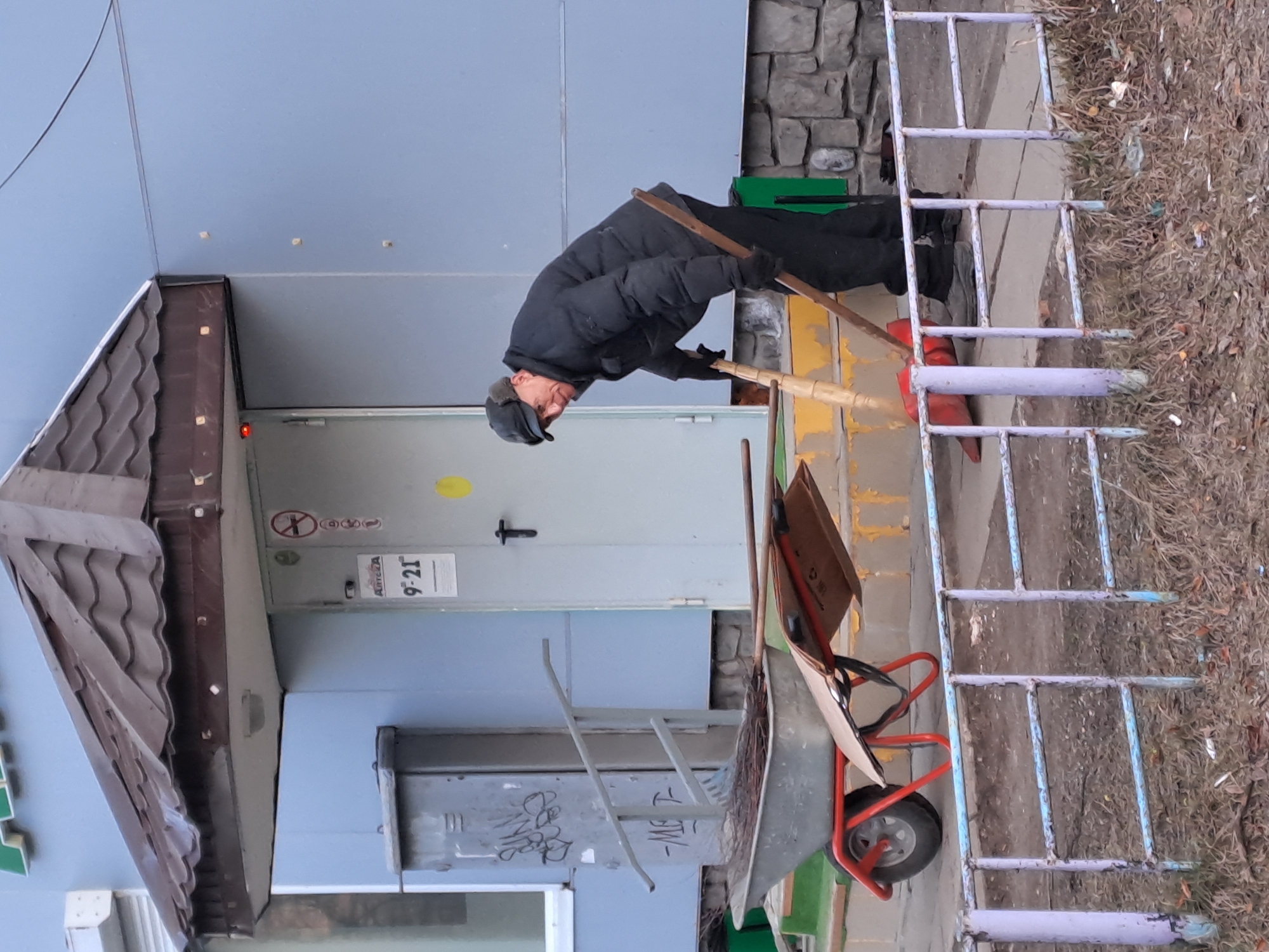 An elderly male, dressed in a black quilted jacket, black trousers, black gloves, and a black flat cap, is observed sweeping debris on the concrete walkway in front of an "APTEKA 9-21" pharmacy building in Elektrostal, Russia. He is positioned center-right, bent forward, actively holding a wooden-handled broom or rake, looking towards the viewer. A red plastic dustpan lies on the ground near his feet. To his left, a metal wheelbarrow with a red frame contains swept organic material and two pieces of cardboard resting on its side. The building features a light blue painted facade, a grey entrance door displaying the pharmacy name and operating hours ("APTEKA 9-21"), a "No Smoking" sign, and a yellow circular sticker. A brown, tiled canopy extends over the entrance, housing a light fixture. The building's foundation consists of grey stone blocks, and yellow-painted concrete steps lead to the door. To the left of the entrance, a grey metal utility box is visible, marked with black graffiti. In the foreground, a low, weathered metal railing, painted in faded blue and purple hues, separates the walkway from a patch of dry grass and scattered organic debris. The scene depicts outdoor cleaning activity in a public urban setting.
