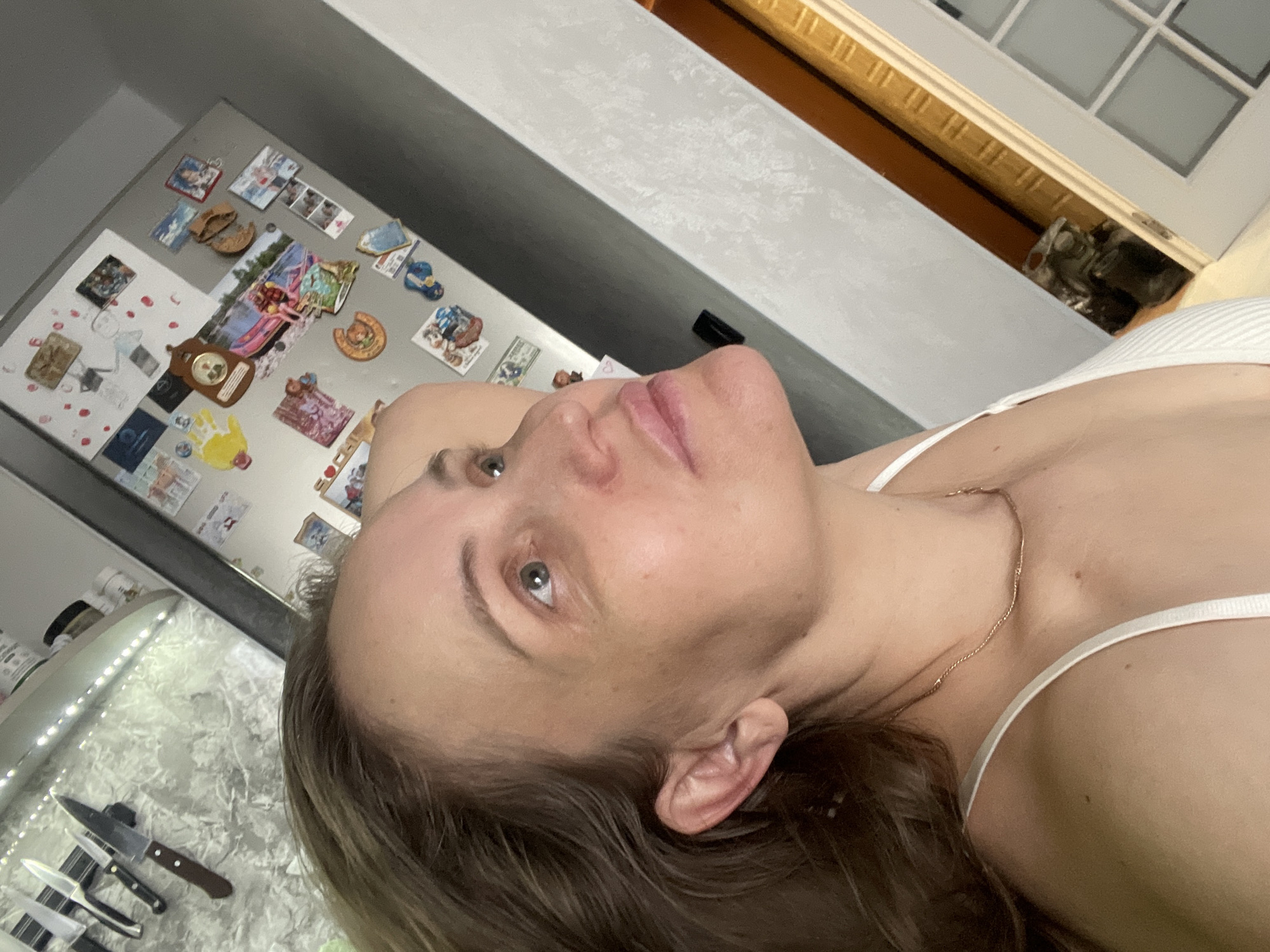 A selfie taken indoors, likely in a kitchen, based on the presence of a refrigerator with magnets, counter and knives. The individual has fair skin, hazel eyes, and brown hair. A gold necklace is visible. The location in the city of Chaikovsky, Russia, cannot be confirmed or denied based solely on the image.