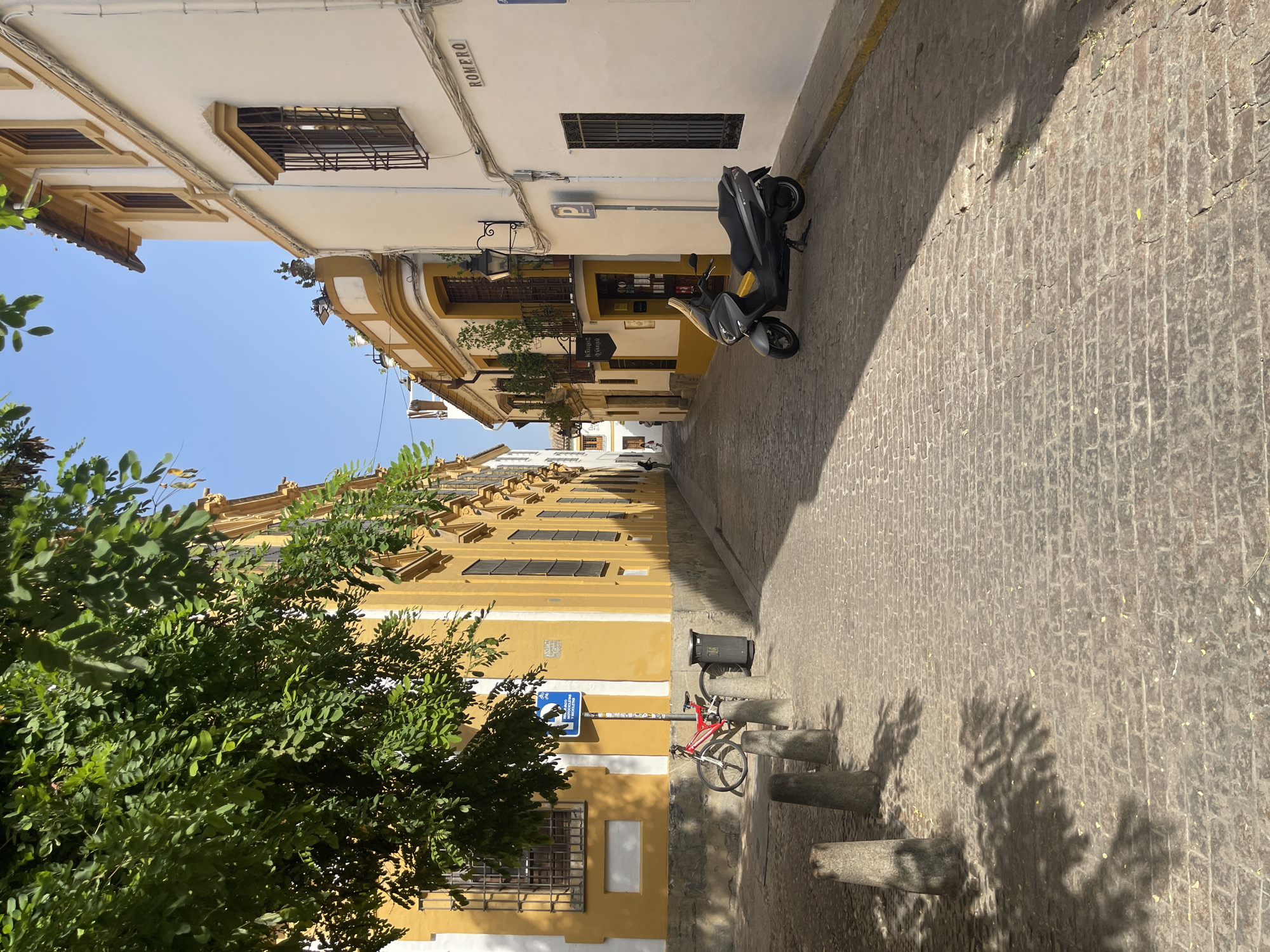 The image shows a narrow, cobblestone street in Córdoba, Spain, lined with traditional Andalusian buildings. The architecture features whitewashed walls with yellow accents and wrought-iron window grilles. Lush green trees with dappled sunlight cast shadows on the street and buildings.
On the left side, a bright yellow building with large windows and a shadowed courtyard is visible. A red bicycle is parked near a trash bin and several stone bollards. A blue parking sign with a white 'P' is mounted on a pole.
On the right side, a white building with yellow trim features a street sign reading "ROMERO." A gray scooter is parked in front of this building, which has a dark awning over its windows. The street is paved with uneven cobblestones, and a distinct shadow line divides the sunlit and shaded portions of the pavement. In the distance, the street curves and narrows, with a few figures walking, suggesting a residential or commercial area. The sky is clear and blue.